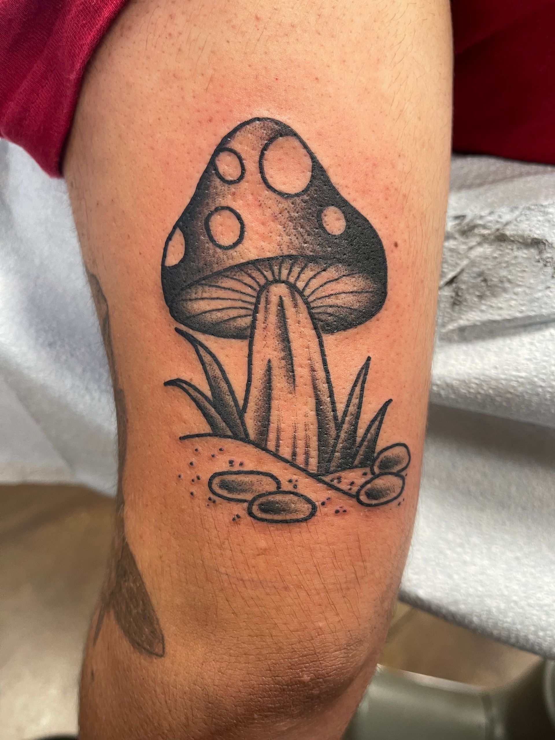 Black and gray mushroom tattoo on an arm, with grass, ground, and dots.