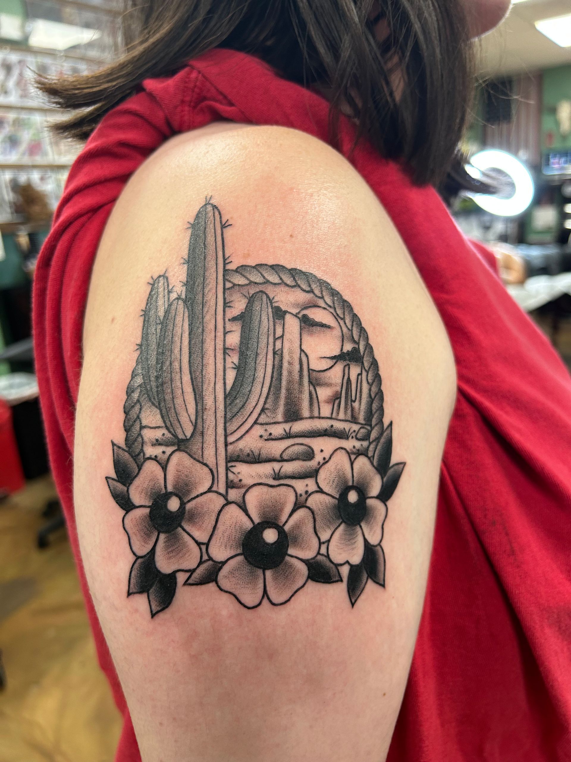 Black and white desert landscape tattoo on a shoulder, featuring a cactus, rocks, and flowers.