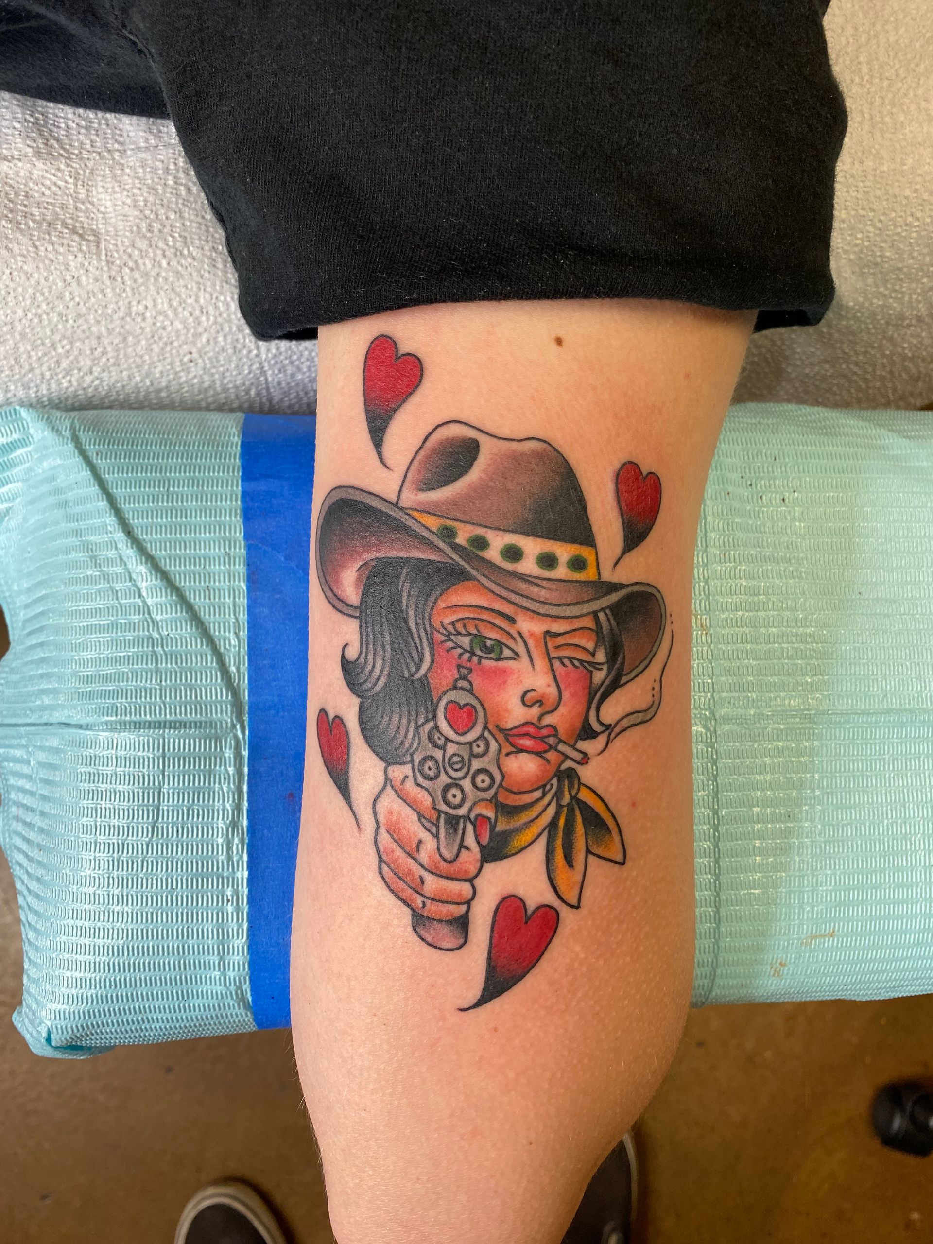 Tattoo of a person in a cowboy hat holding a gun, surrounded by red hearts, on an arm.