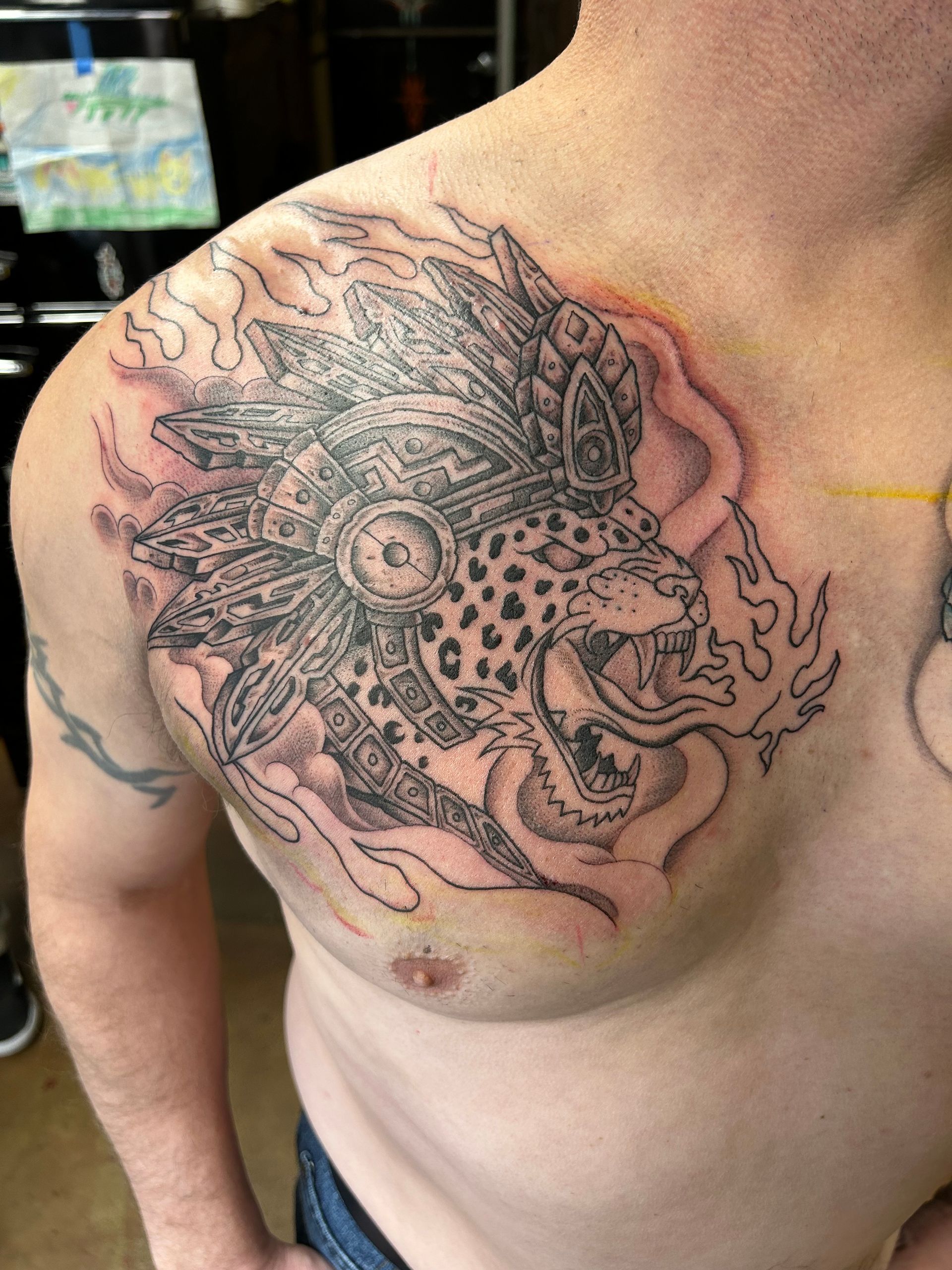 Black ink jaguar with a headdress tattoo on a person's chest, surrounded by smoke/flames.