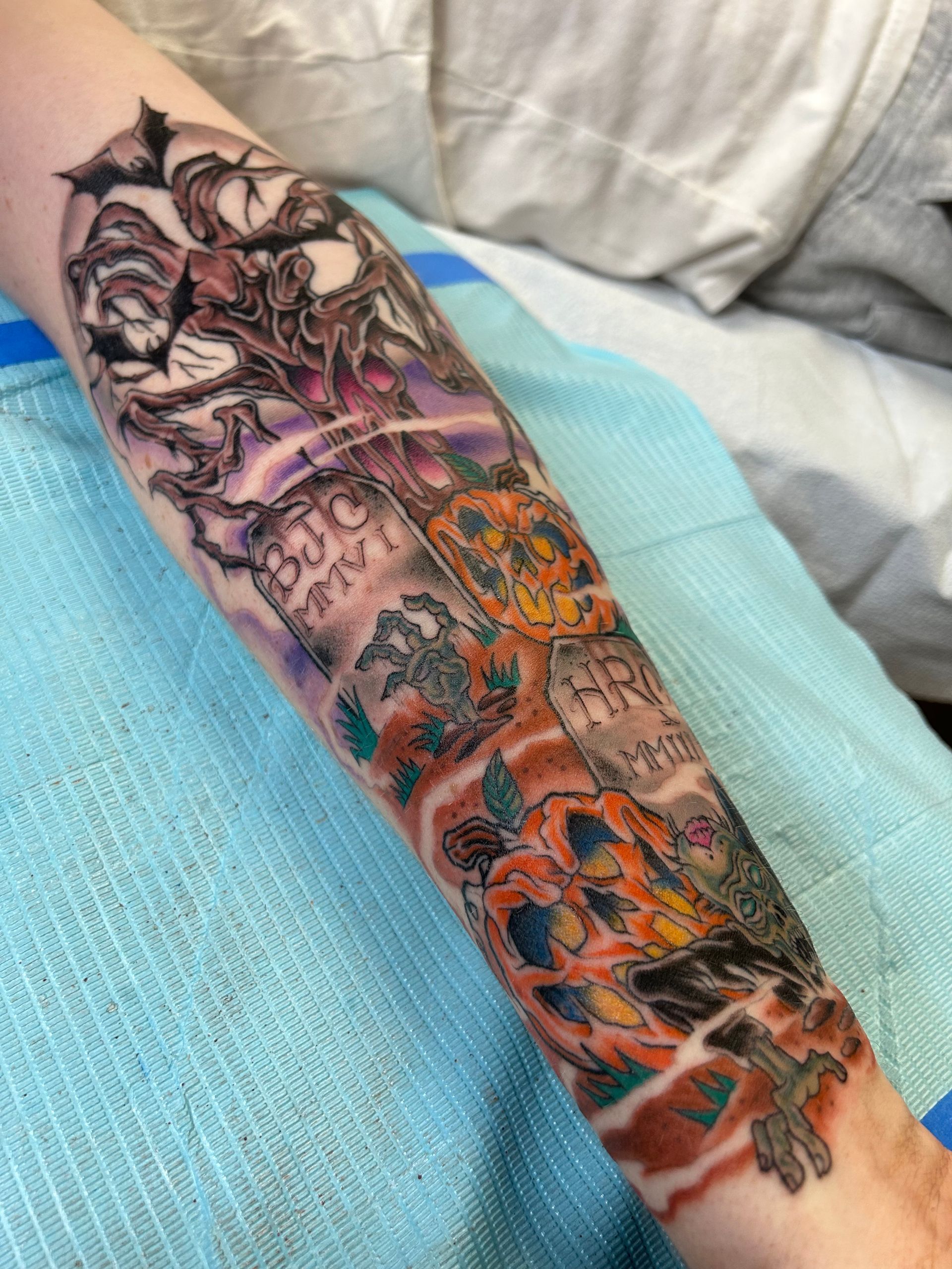 Tattoo on arm depicting a graveyard scene with tombstones, orange pumpkins, bats, and flowers, in various colors.
