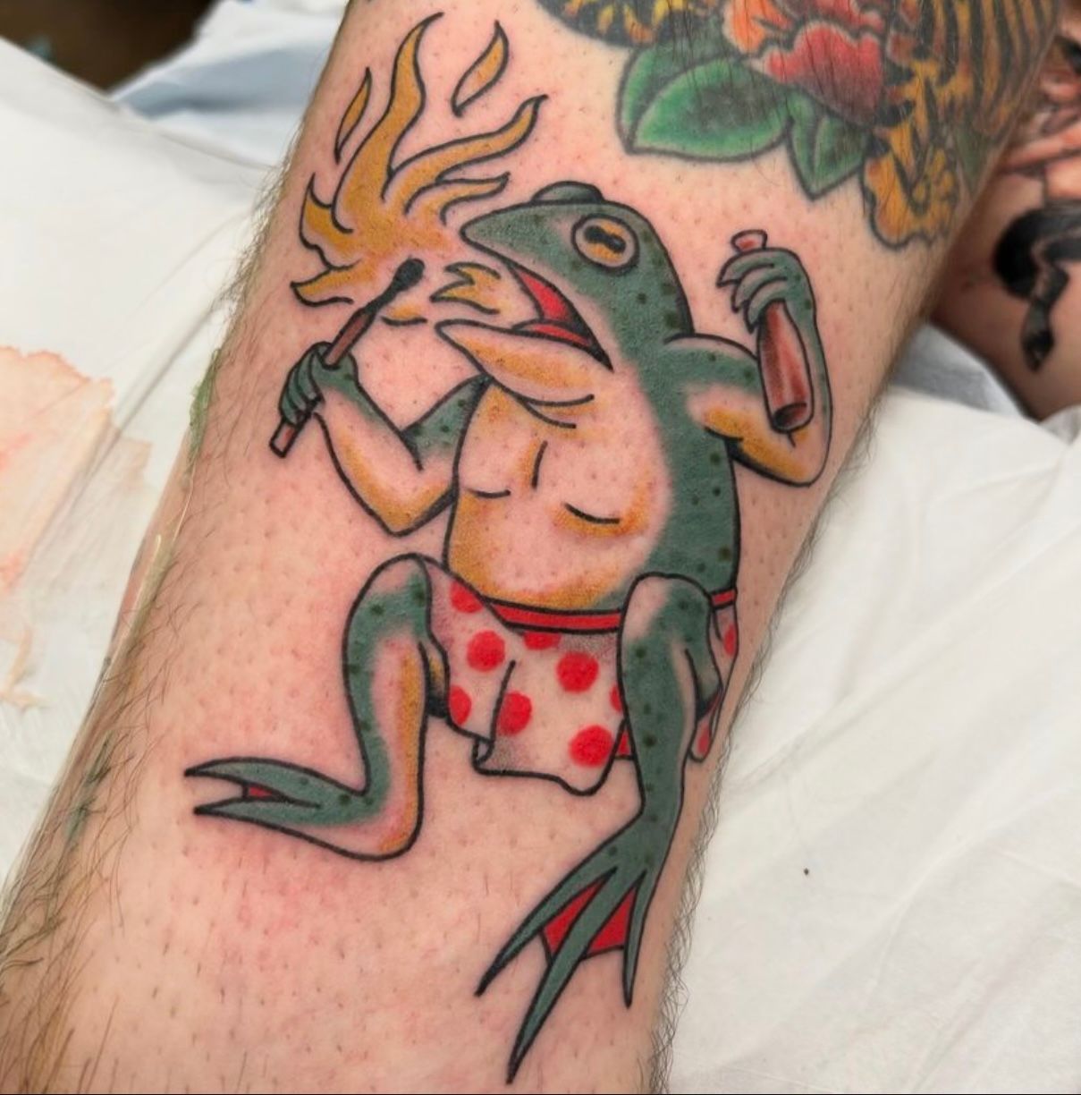 Tattoo of a frog holding a lit match and beer bottle, wearing red polka-dot shorts.
