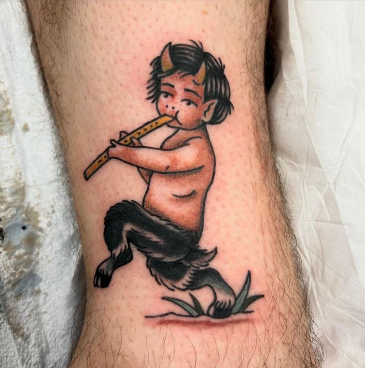Tattoo of a satyr playing a flute. Dark hair, horns, and goat legs.  Red, black, and brown ink on skin.