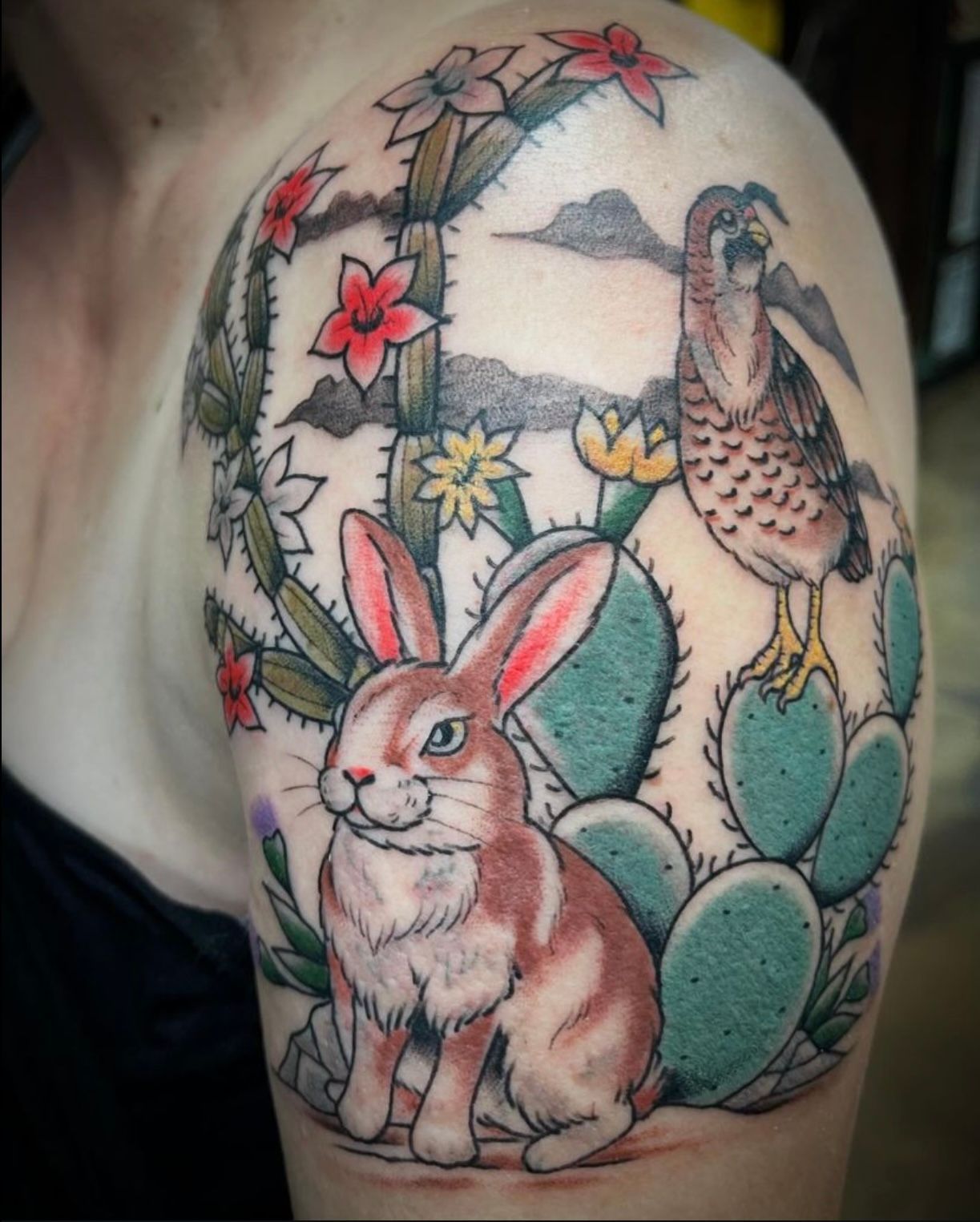 Tattoo of a rabbit, cactus, and bird with a desert theme in shades of green, red, and brown on a shoulder.
