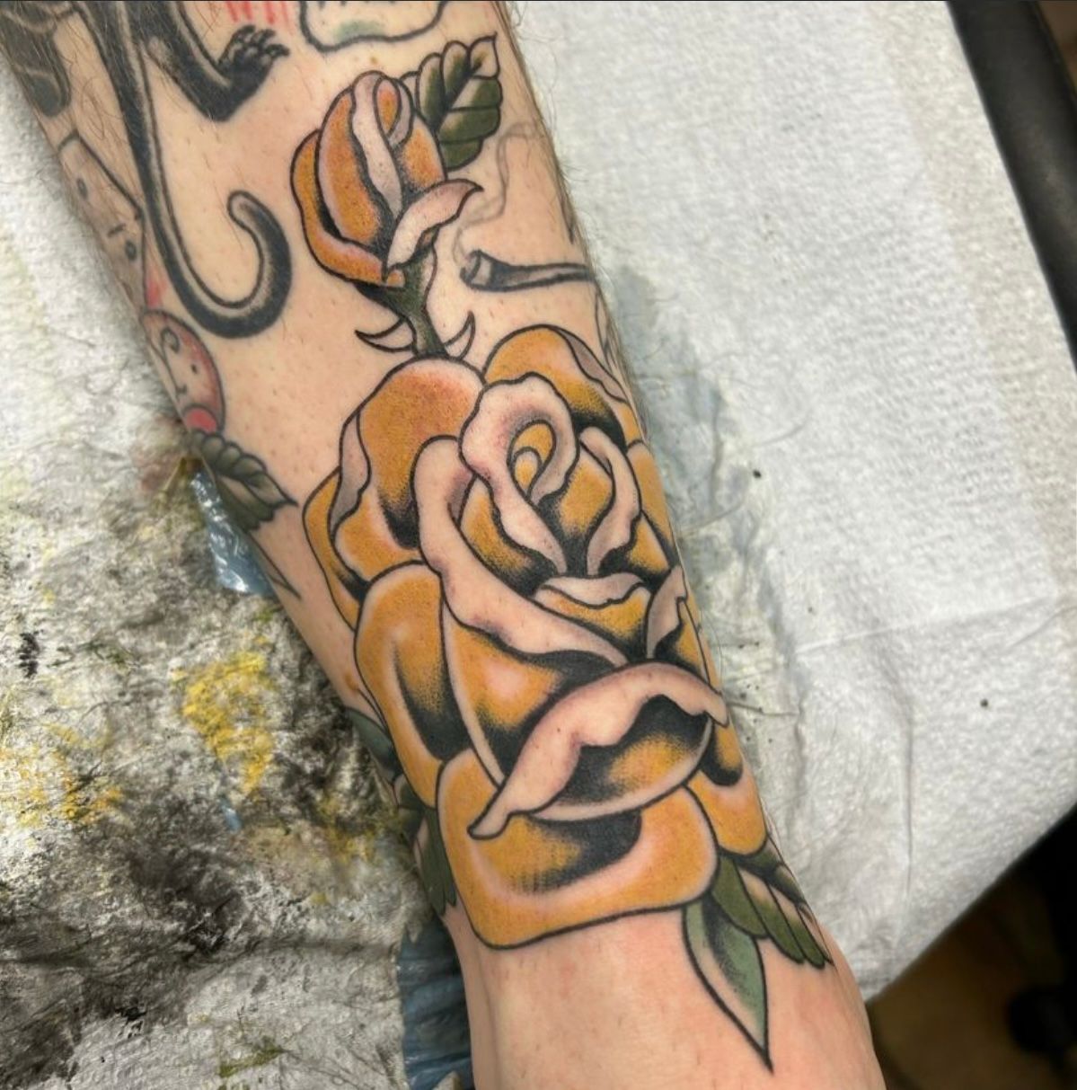 Yellow rose tattoo on a forearm. Black outlines, green leaves, and black shading.
