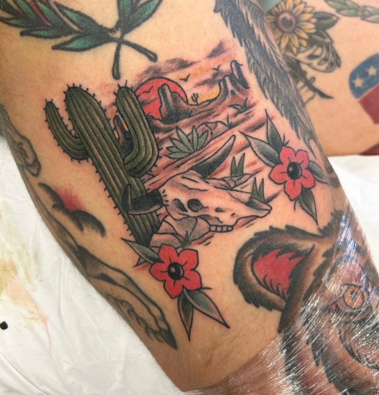 Tattoo of a desert landscape with cactus, dinosaur skull, red flowers, and setting sun.