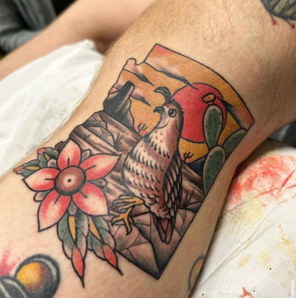 Tattoo: bird perched on branch in a desert scene; red sun, cactus, flower, brown, orange.