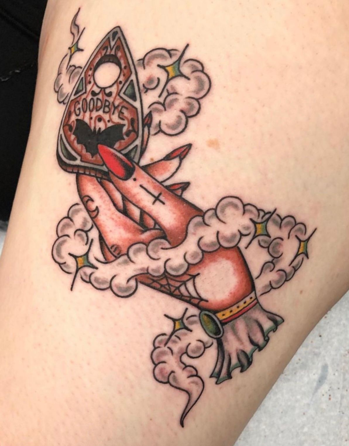 Tattoo of a hand holding a Ouija planchette, surrounded by smoke and stars.