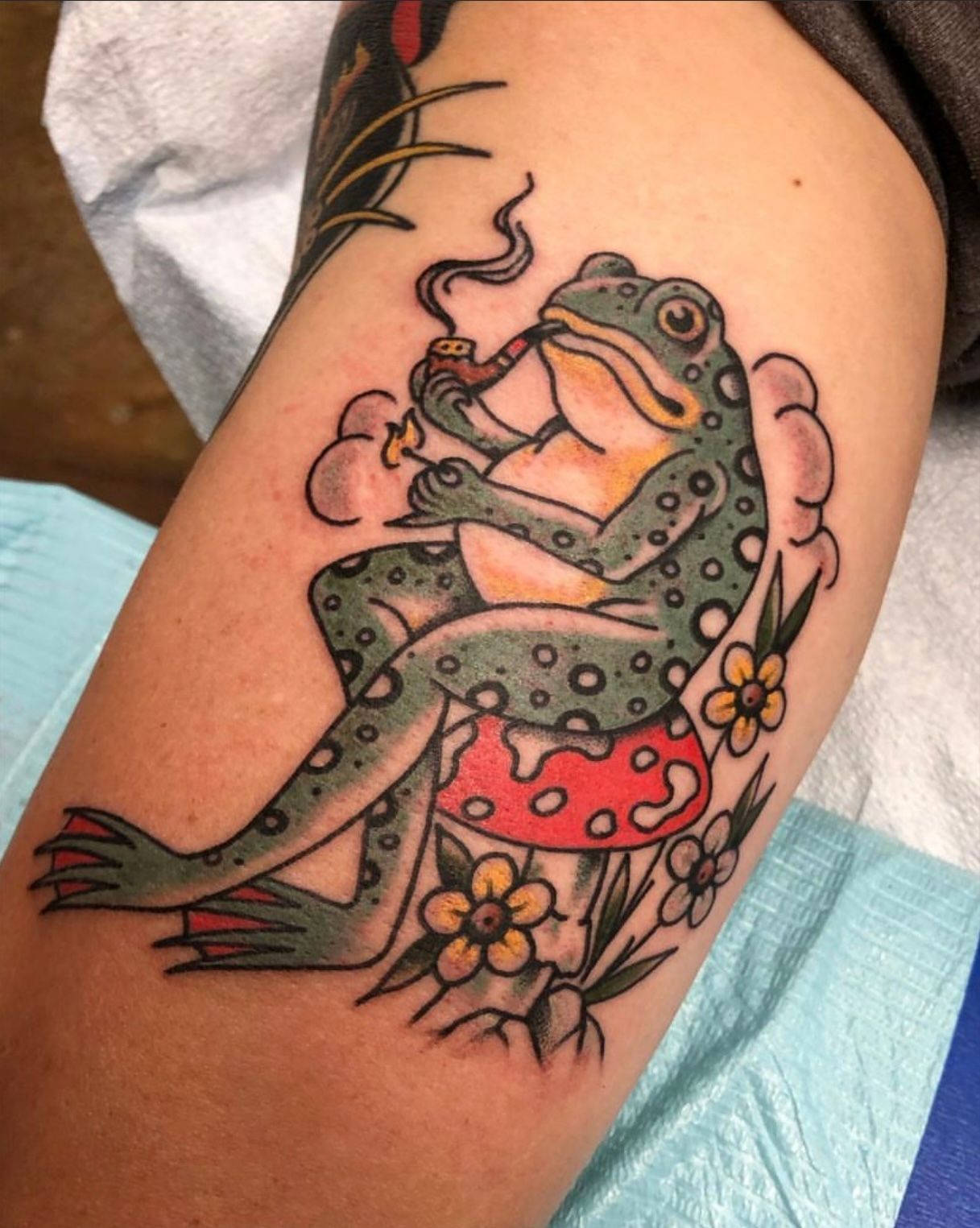 Tattoo of a frog smoking a pipe, sitting on a mushroom. Green frog, red mushroom, yellow flowers, and smoke.