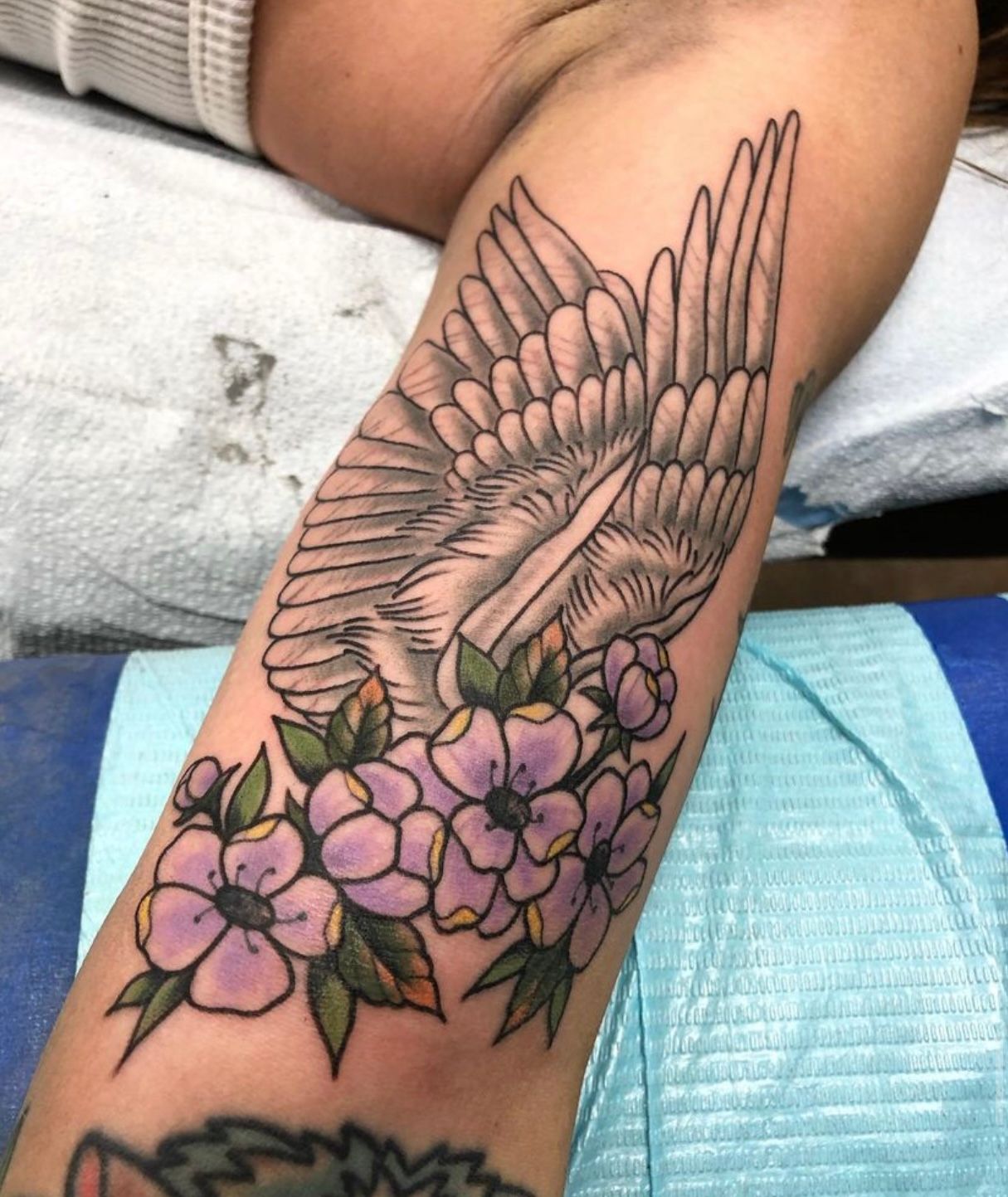 Tattoo of a white wing above purple flowers on a person's arm, shaded details.