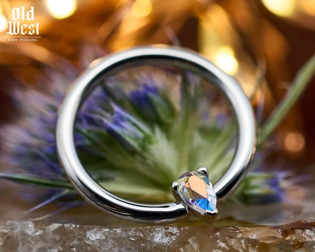 Silver hoop earring with a teardrop-shaped iridescent gem, resting on a textured surface with blurred floral background.