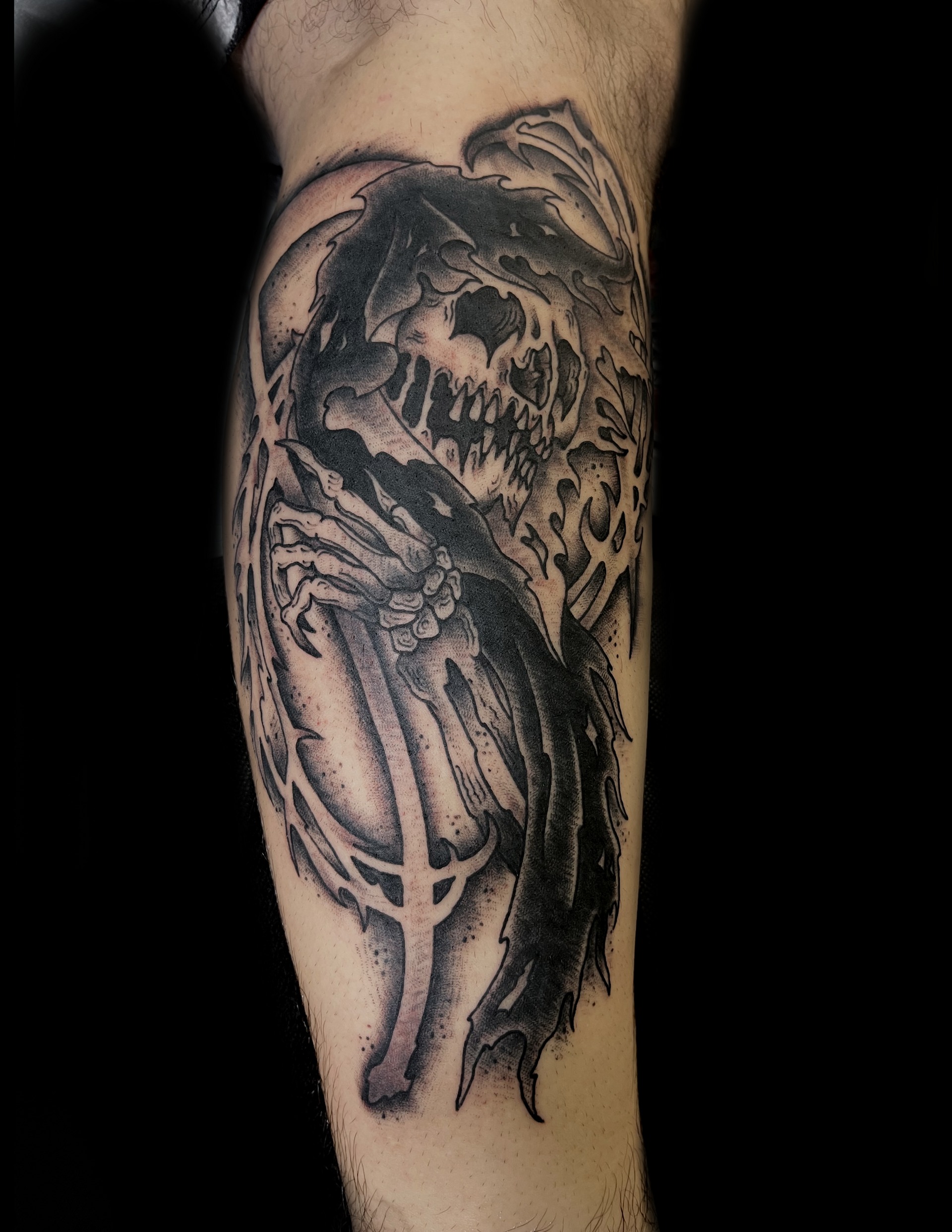 Tattoo of a skull and skeletal arms holding an inverted cross on an arm, black and gray shading.