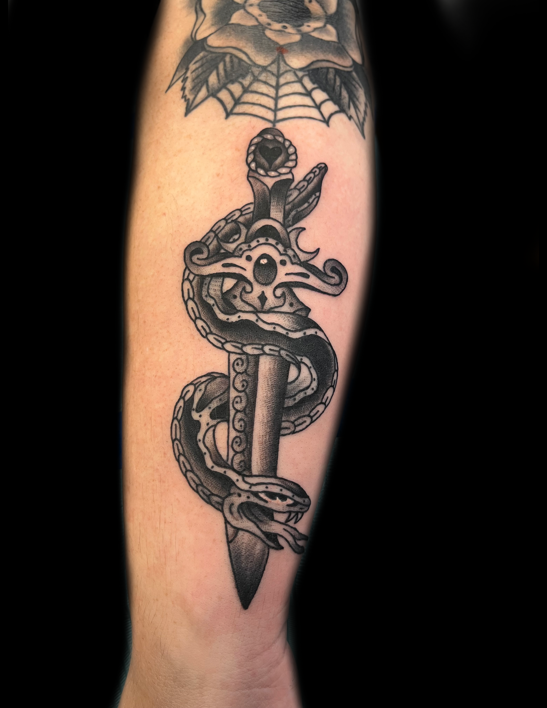 Tattoo of a dagger with a snake wrapped around it on an arm. Above, a spiderweb design.