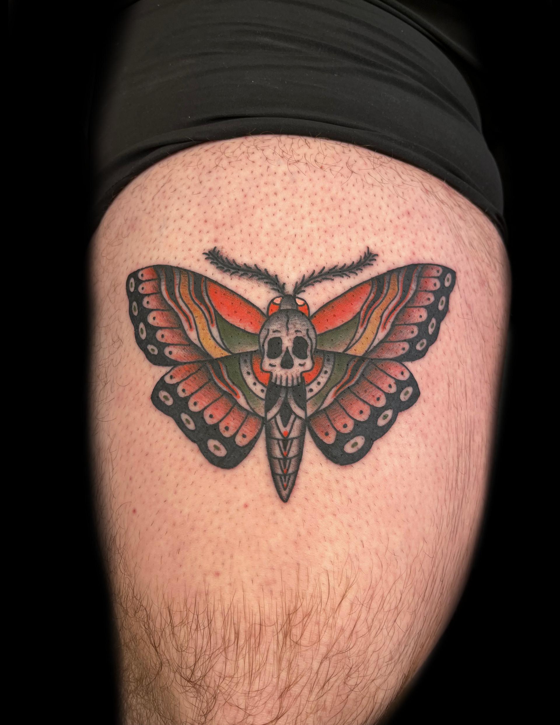 Tattoo of a moth with a skull on a leg. Red, black, and green wings with detailed linework.