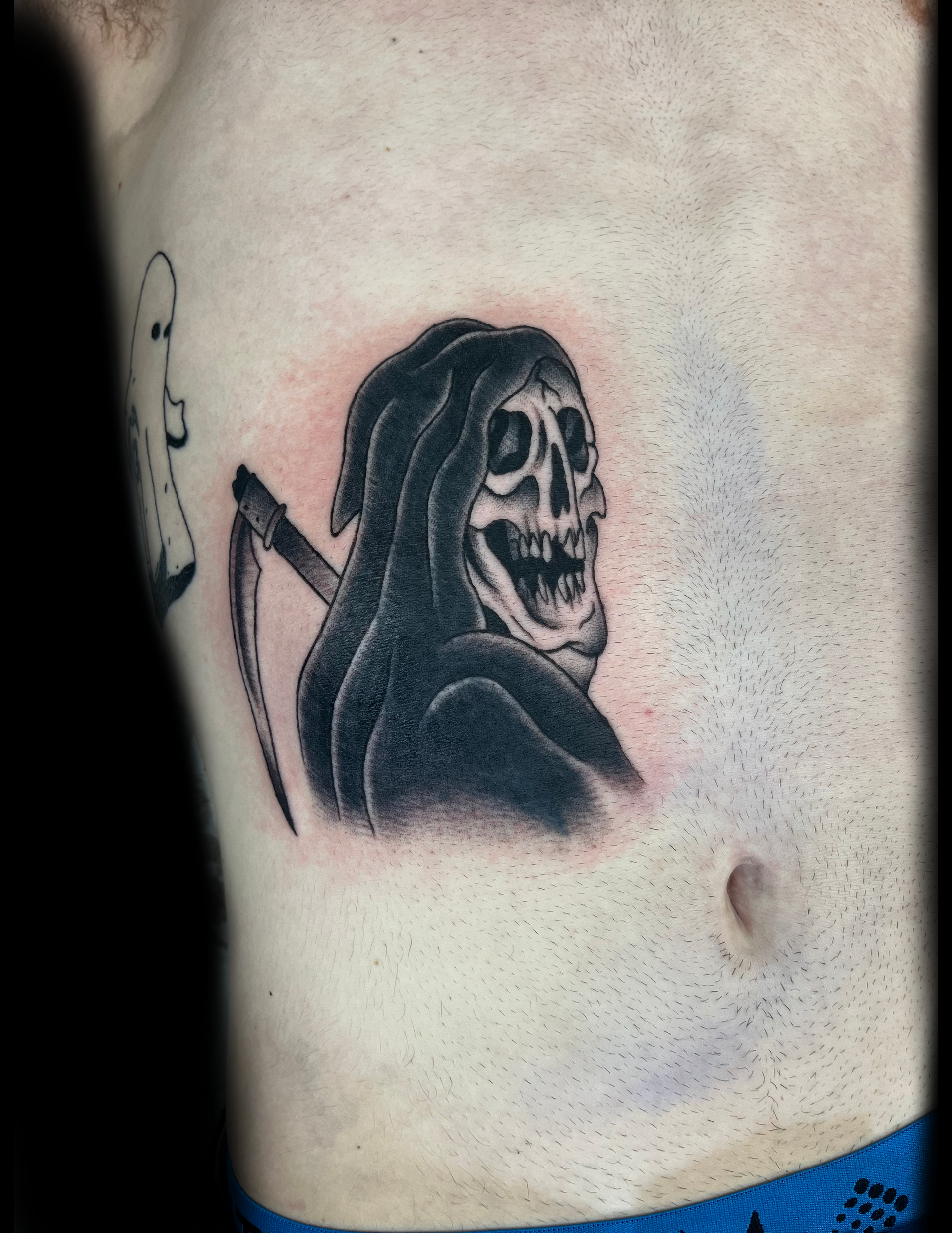 Tattoo of a grim reaper skull with scythe on a person's torso; black ink on skin.