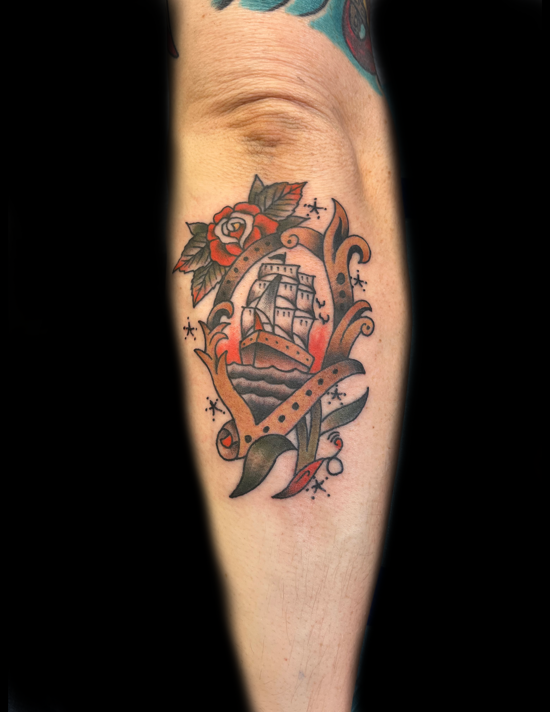 Tattoo on arm; ship inside a frame with a rose, red and gold details.