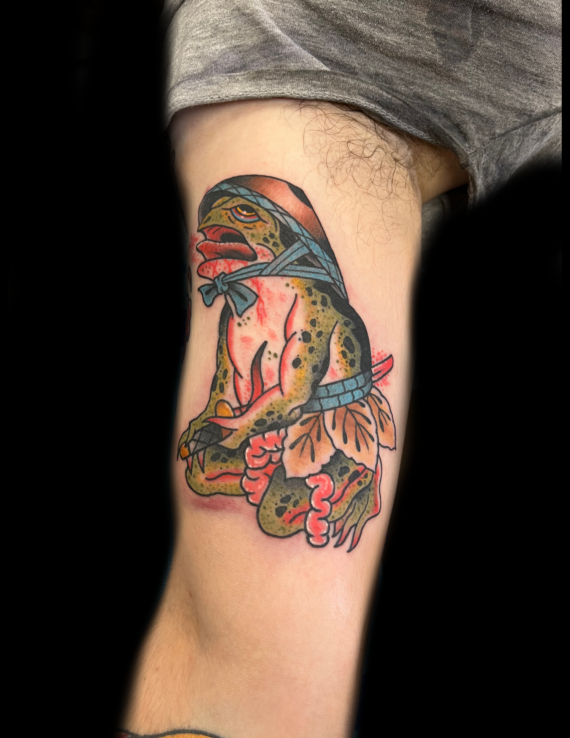 Tattoo of a green frog in a headscarf and flower skirt, seated with hands clasped; arm.