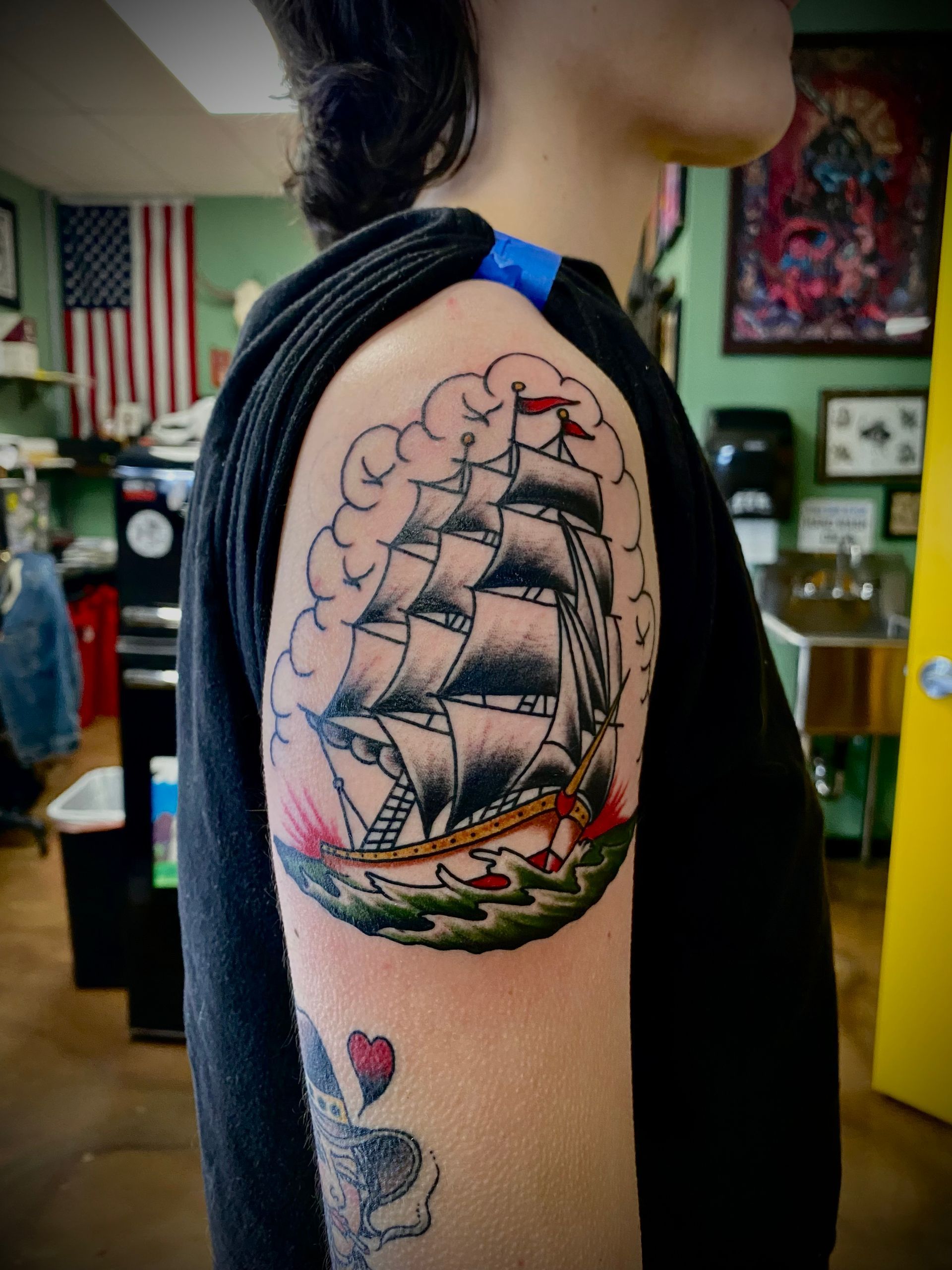 Tattoo of a ship sailing through stormy waters on a person's upper arm. Clouds, waves, and red accents.