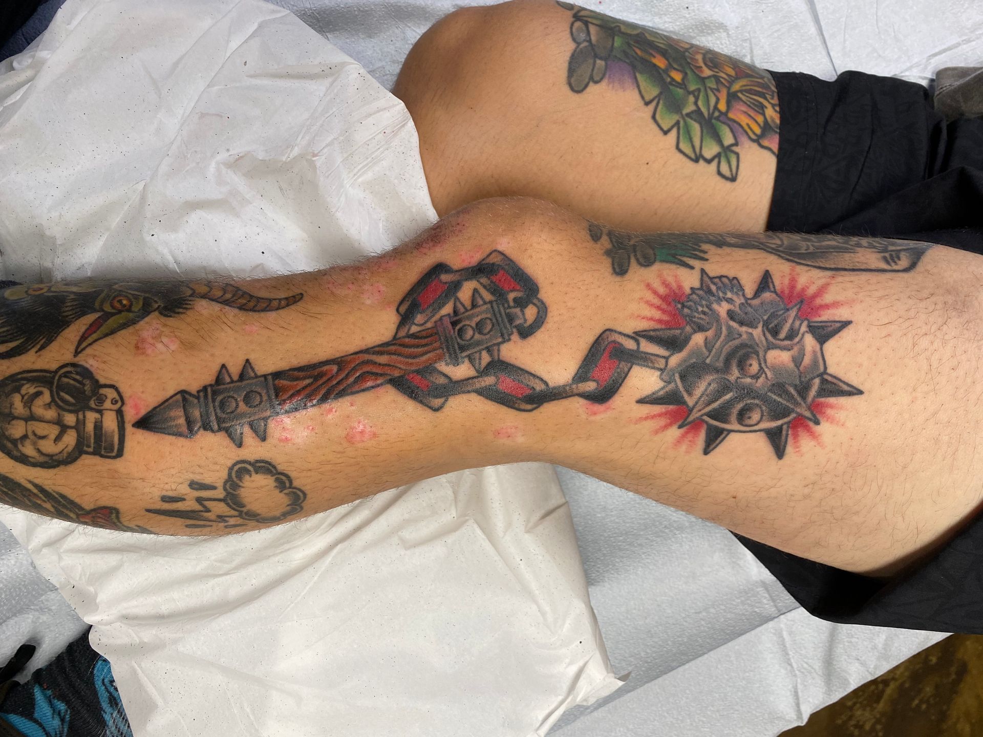 Tattoo of a flail on a person's leg with red, black, and gray ink. Other tattoos are visible.