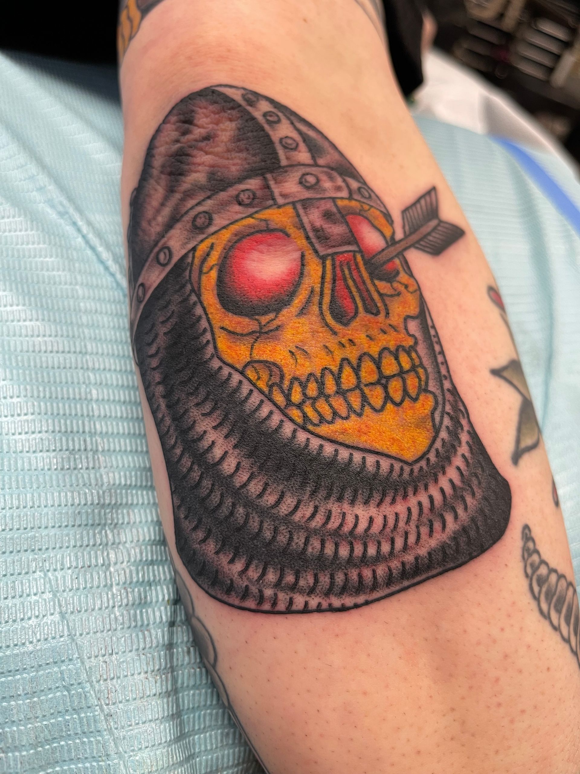 Tattoo of a yellow skull in a helmet with an arrow through the eye socket, on an arm.