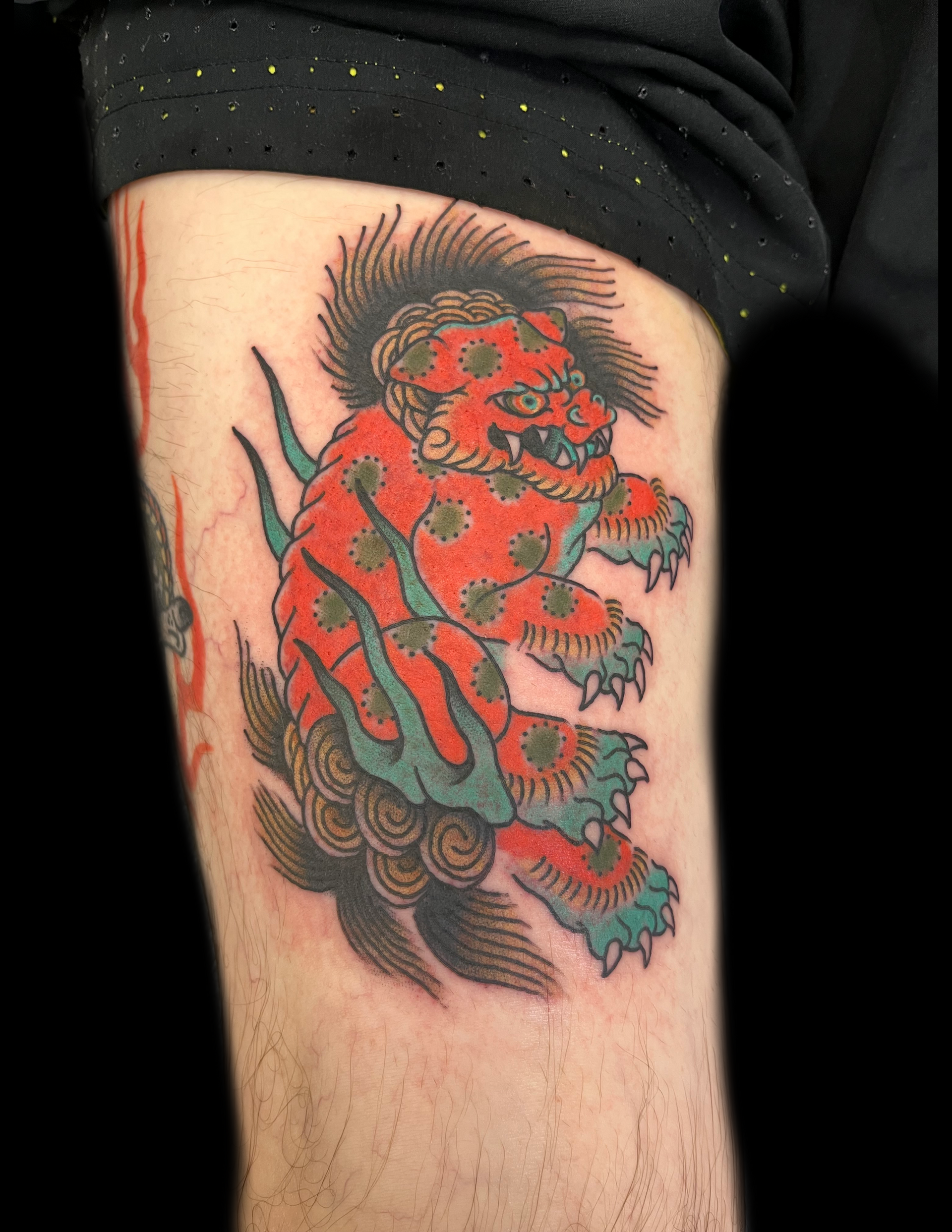 Red and blue Japanese Foo Dog tattoo on thigh, surrounded by flames and clouds.
