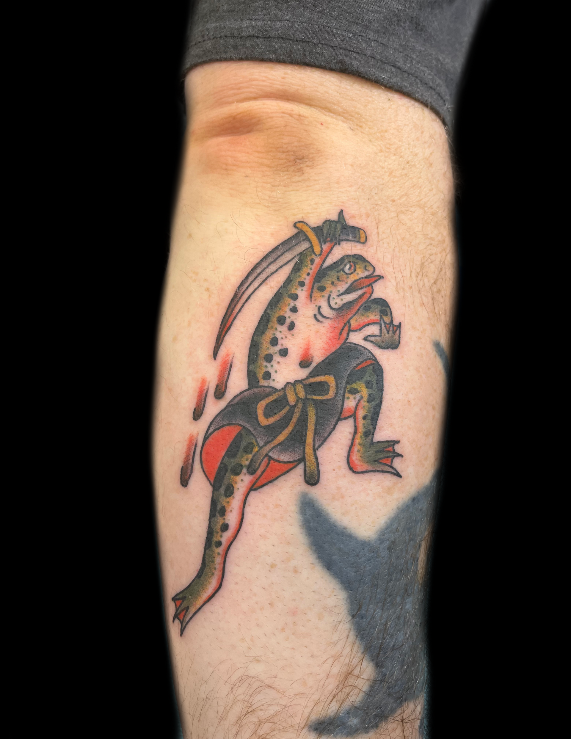 Tattoo of a frog holding a sword, stabbing another frog, with blood drops.
