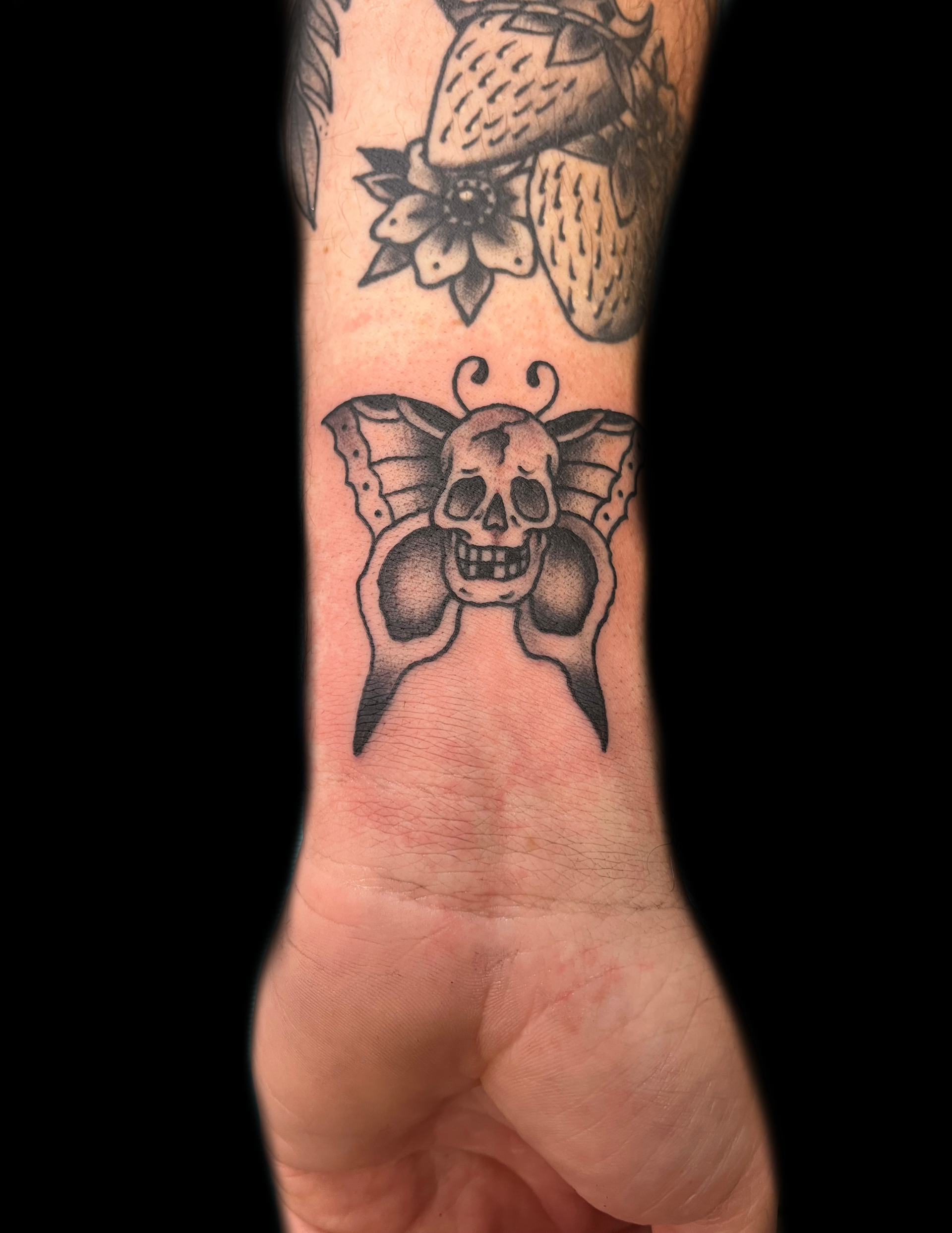 Tattoo on wrist: skull with butterfly wings. Additional tattoos on arm, against a black background.