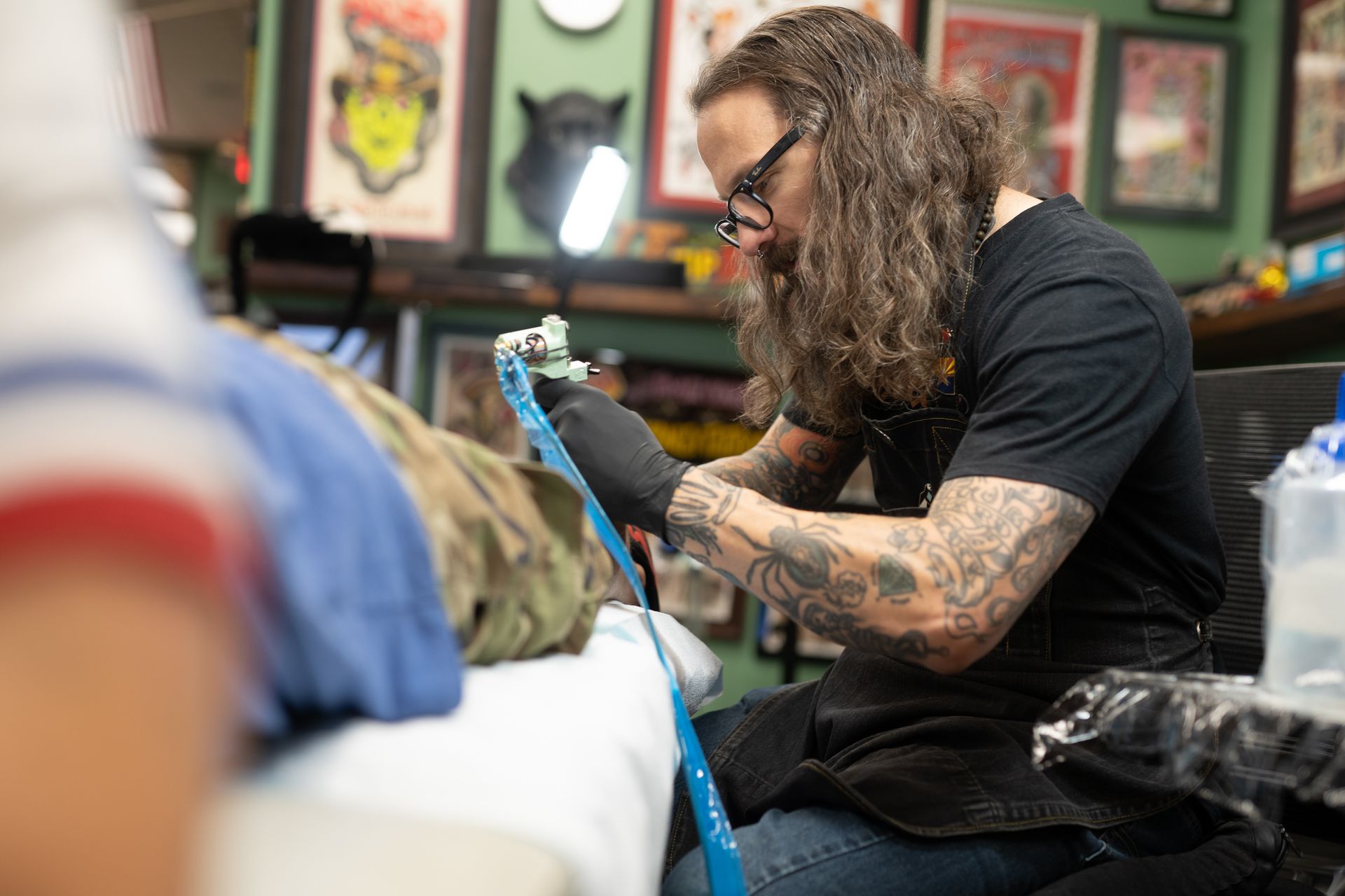 Tattoo artist with tattoos on arms, working on a tattoo. The person wears glasses and a black shirt in a tattoo parlor.
