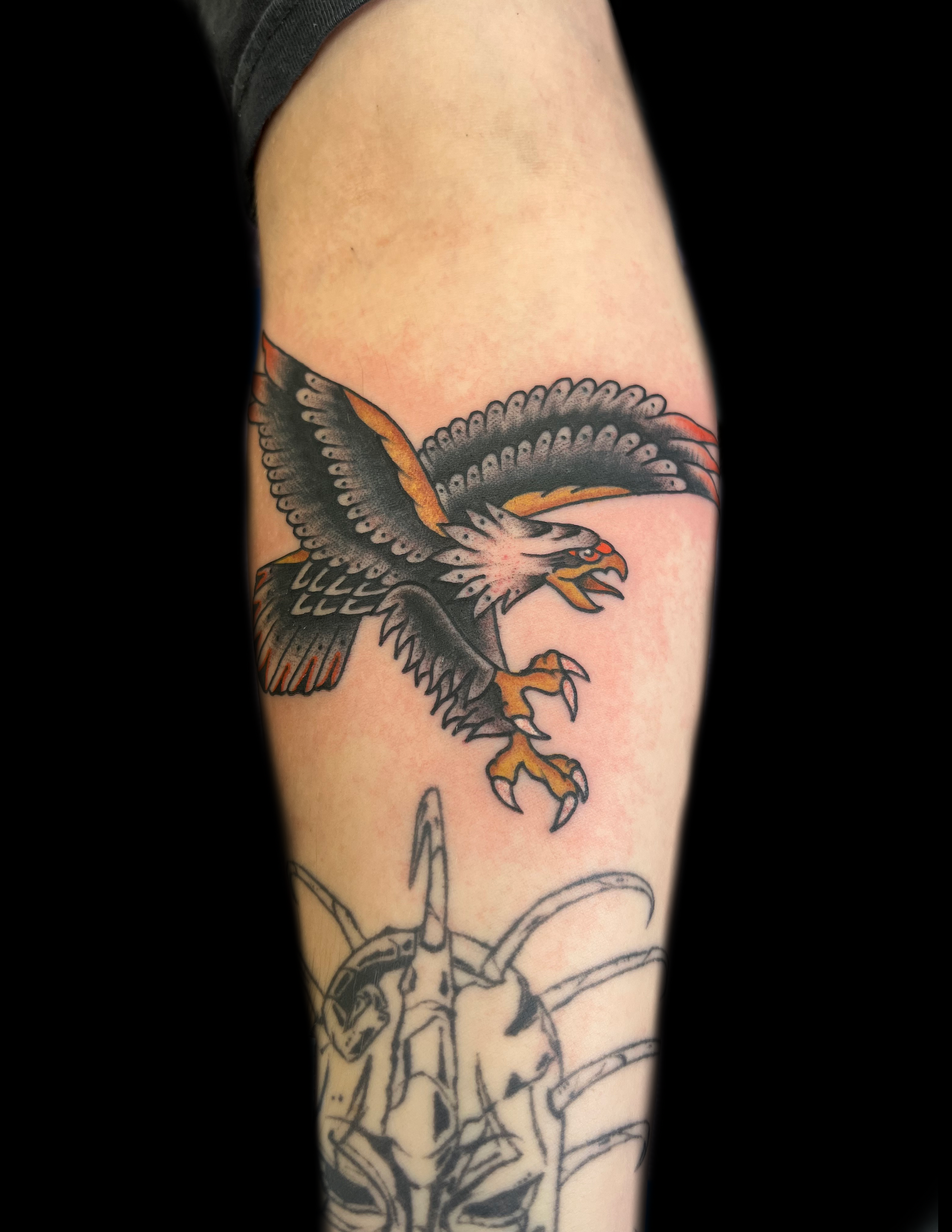 Tattoo of a flying eagle on a person's arm, with wings spread, and claws extended; black, gray, orange.