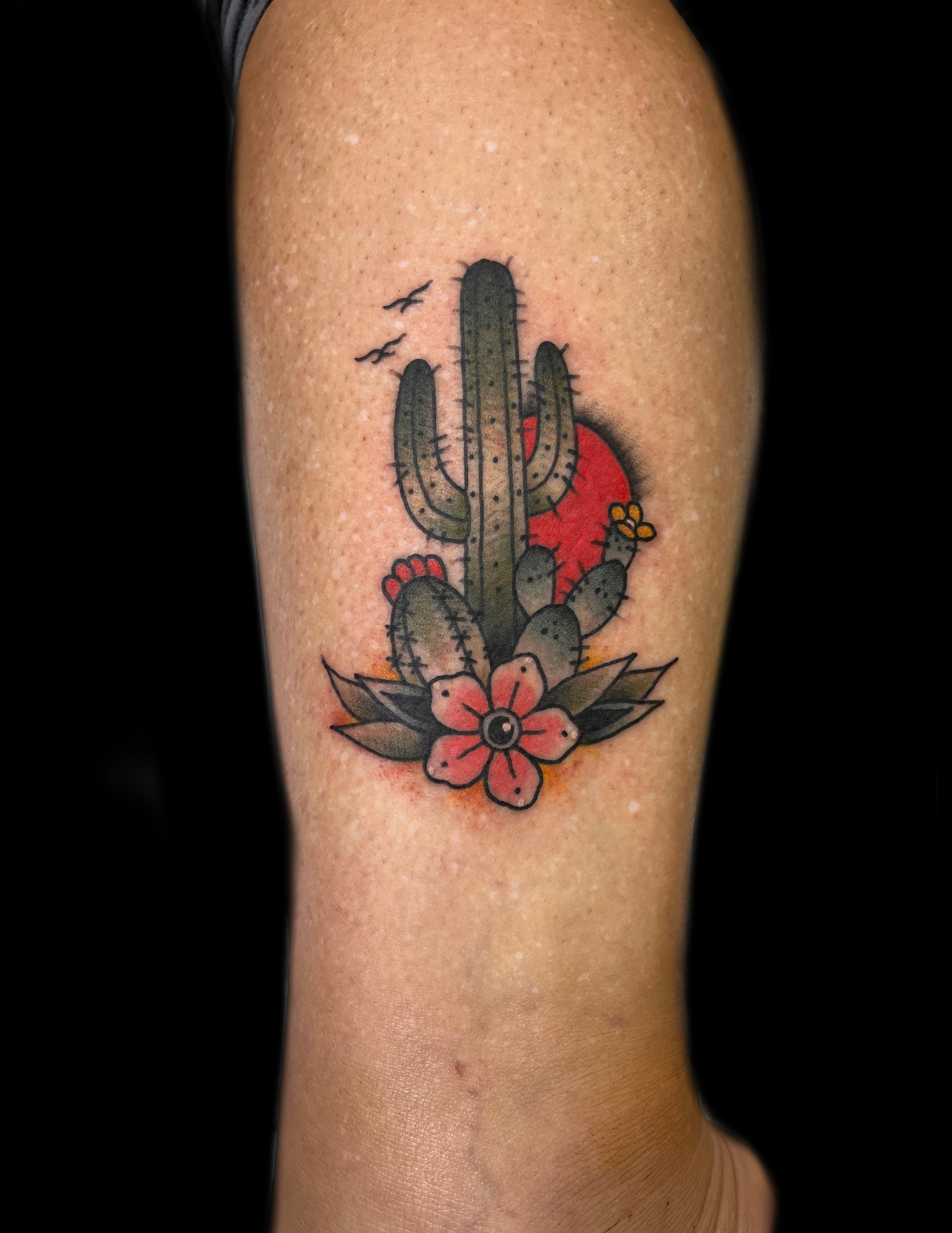 Tattoo of a cactus with a red sun, birds, and flowers on a person's leg.