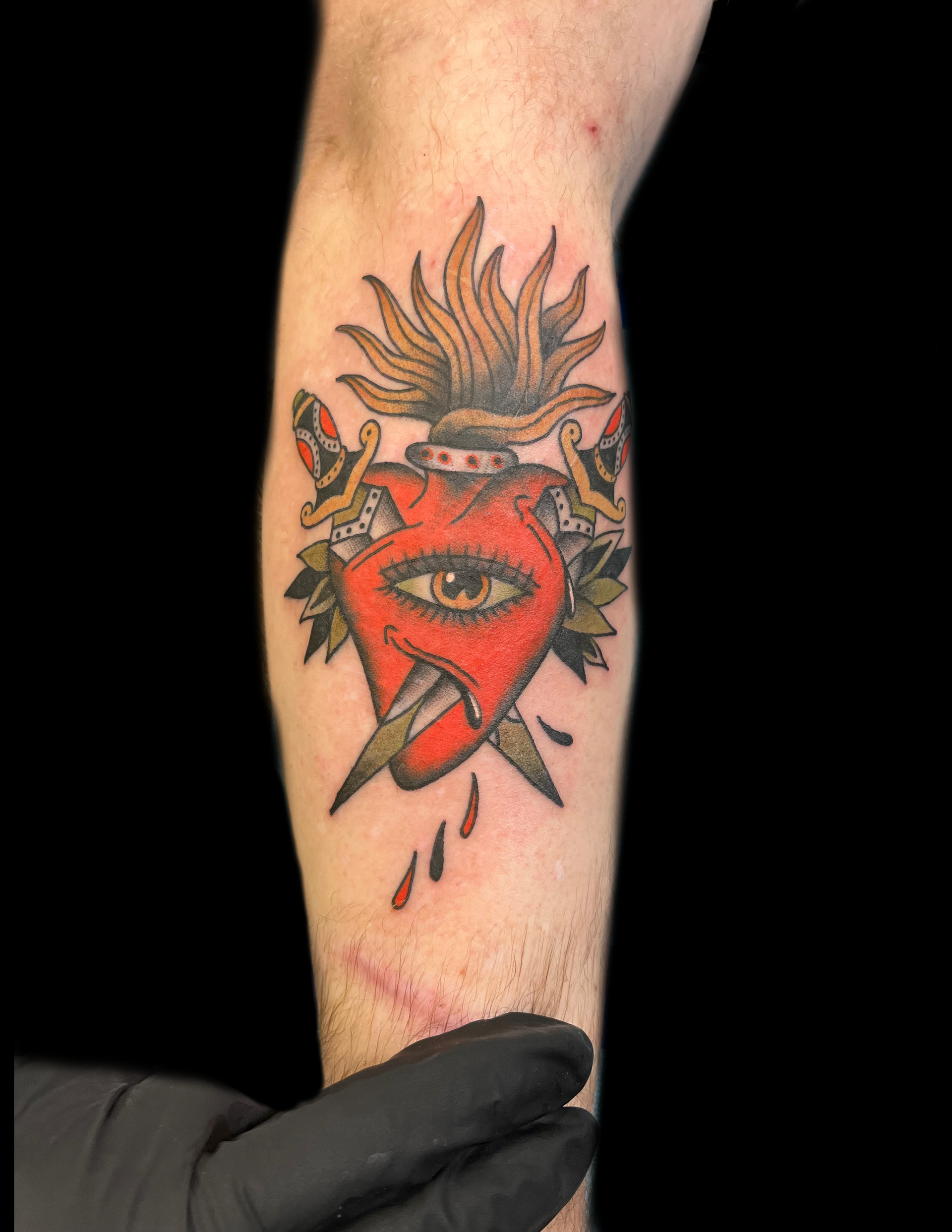 Tattoo of a red heart with an eye, pierced by a dagger, flanked by swords, and topped with flames on an arm.