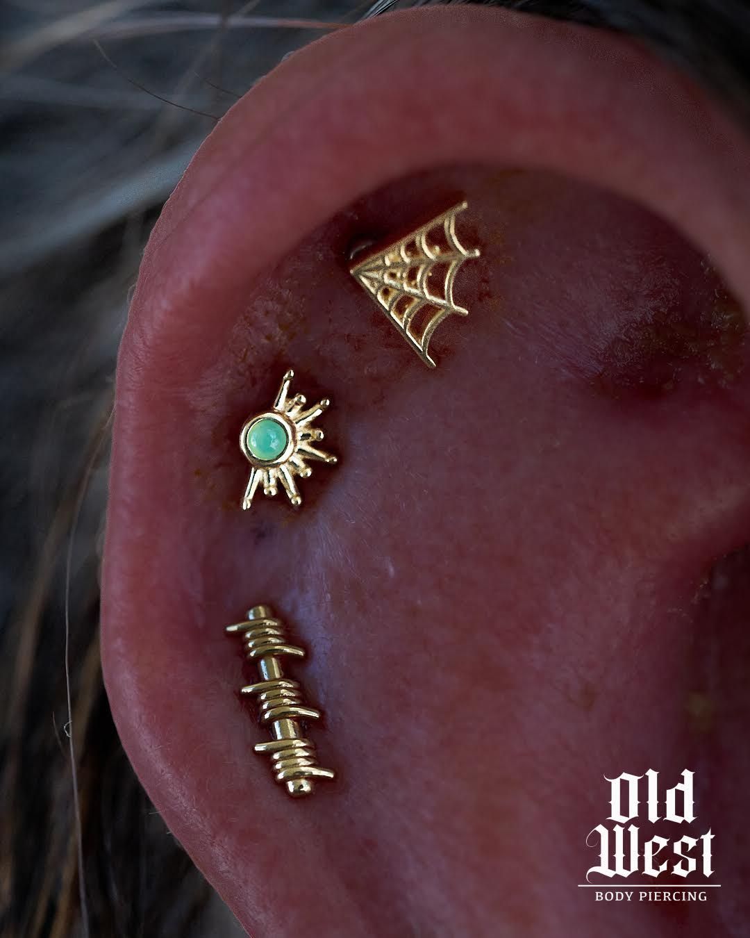 Close-up of an ear with three gold piercing studs: spiderweb, sunburst, and barbed wire.