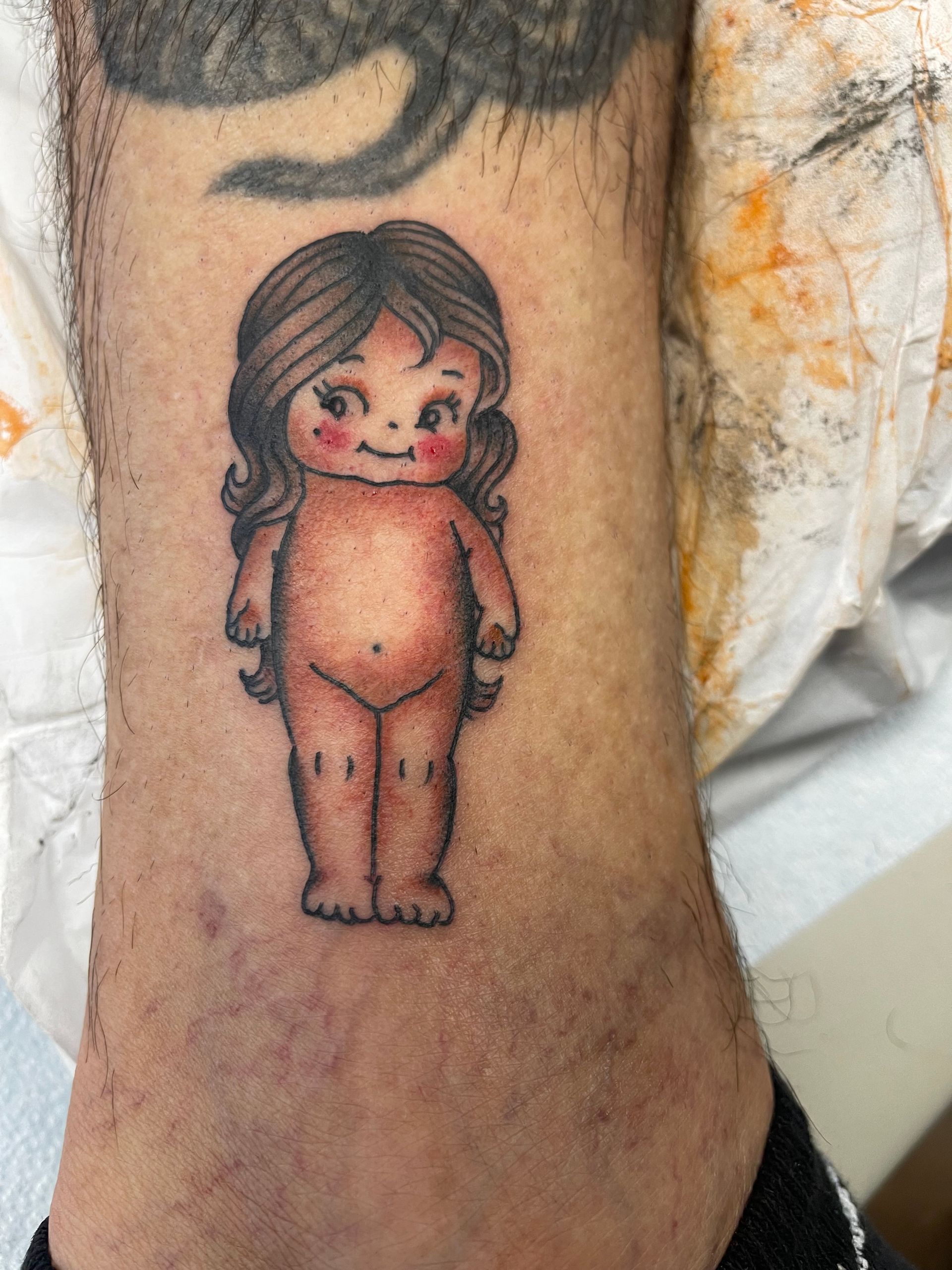 Tattoo of a nude doll with rosy cheeks, brown hair, and black outlines on a leg.