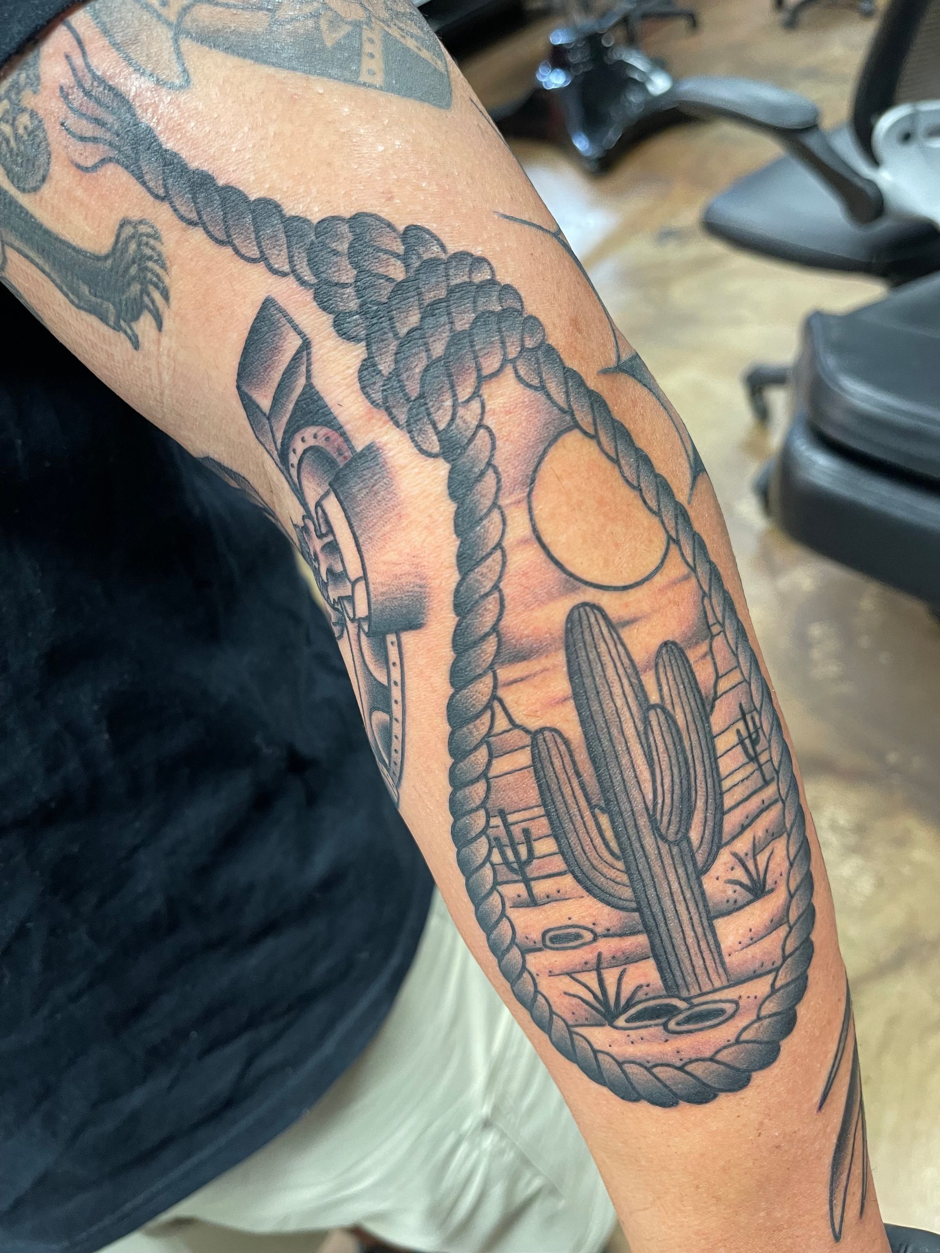 Tattoo of a desert scene within a noose shape on an arm. Features a cactus, sun, and rope detailing.