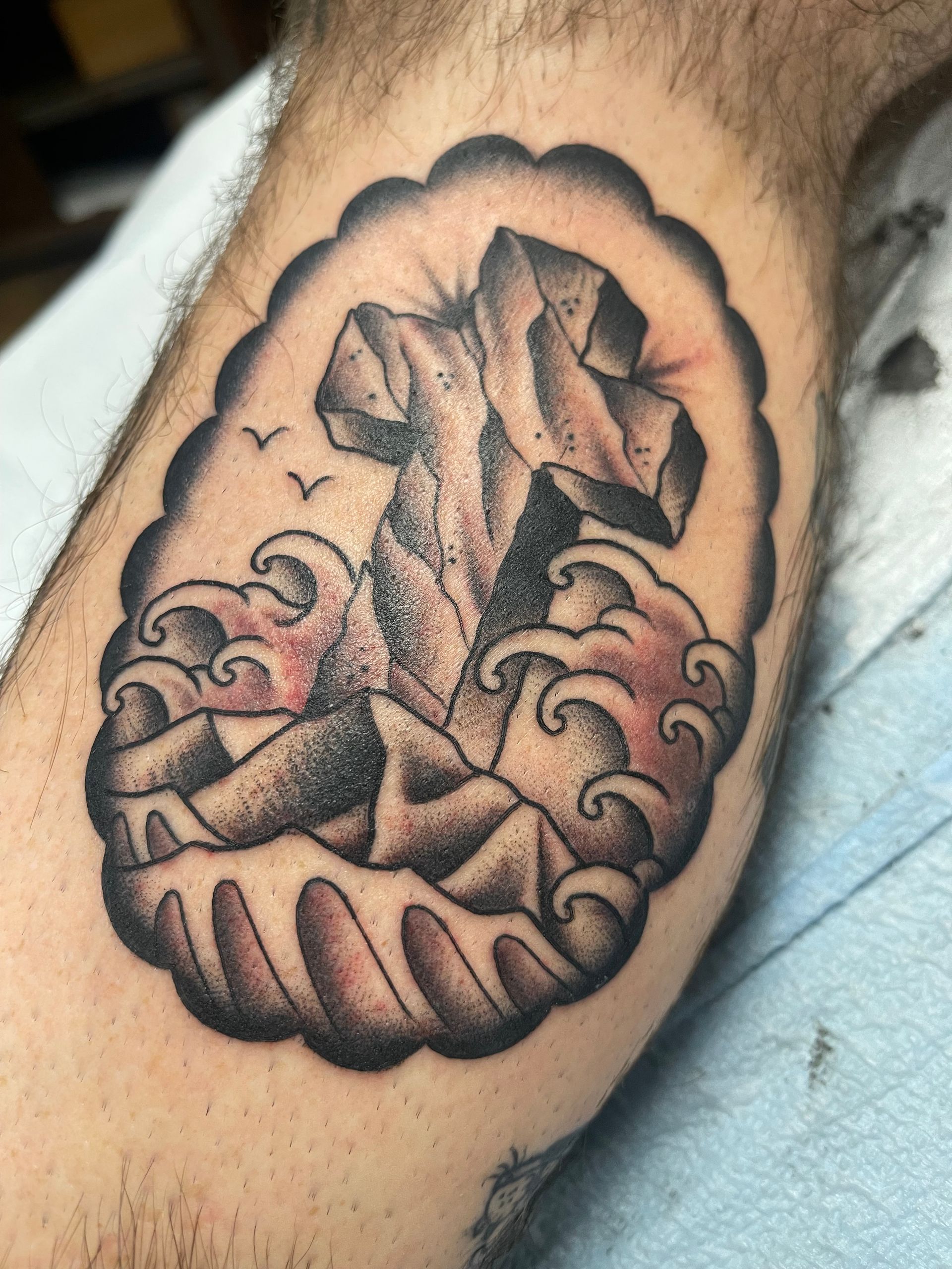 Tattoo of a stone cross emerging from waves, framed in a scalloped border. Two birds fly overhead.