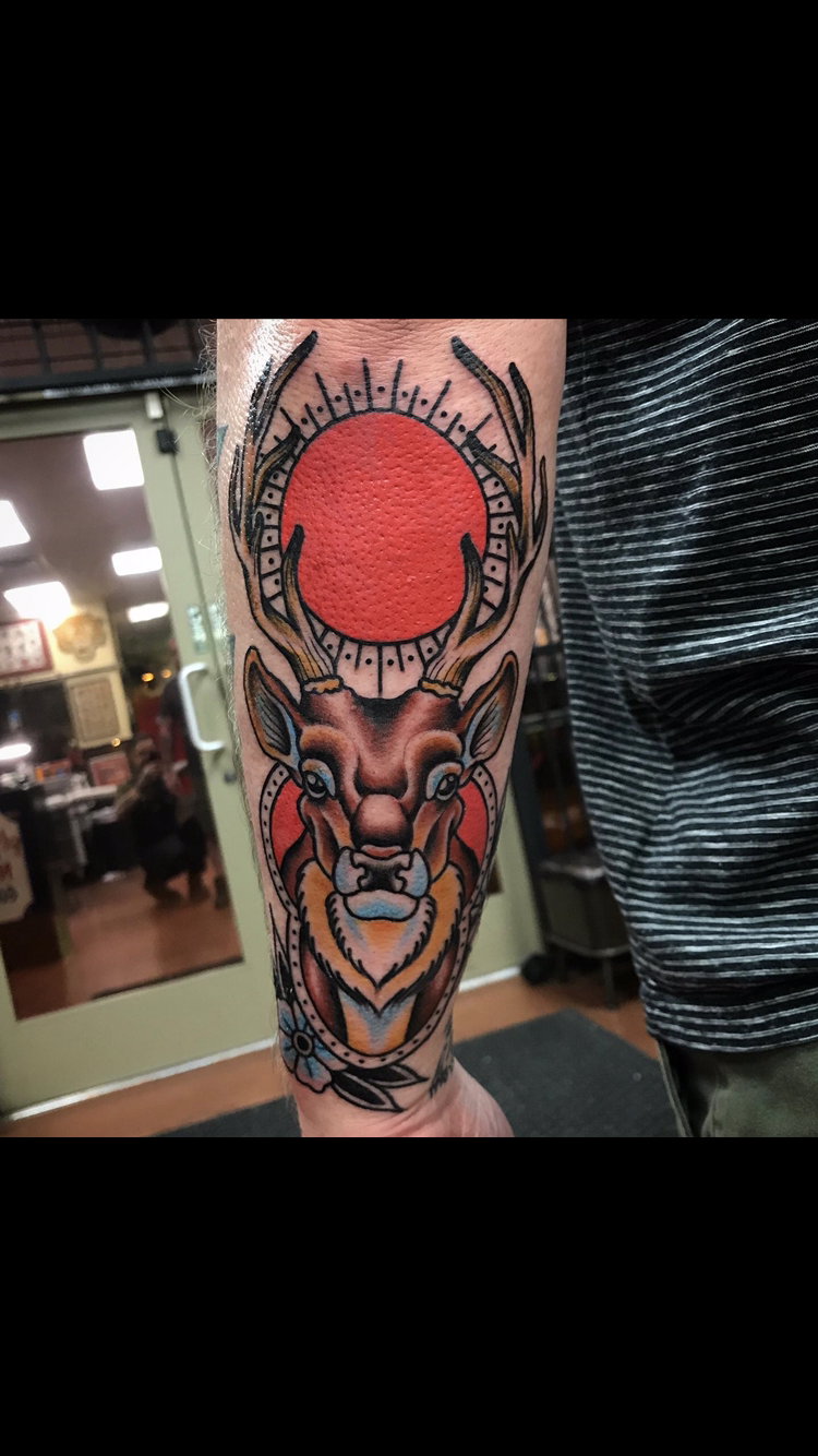 Tattoo of a deer head with large antlers and a red sun, on a forearm.