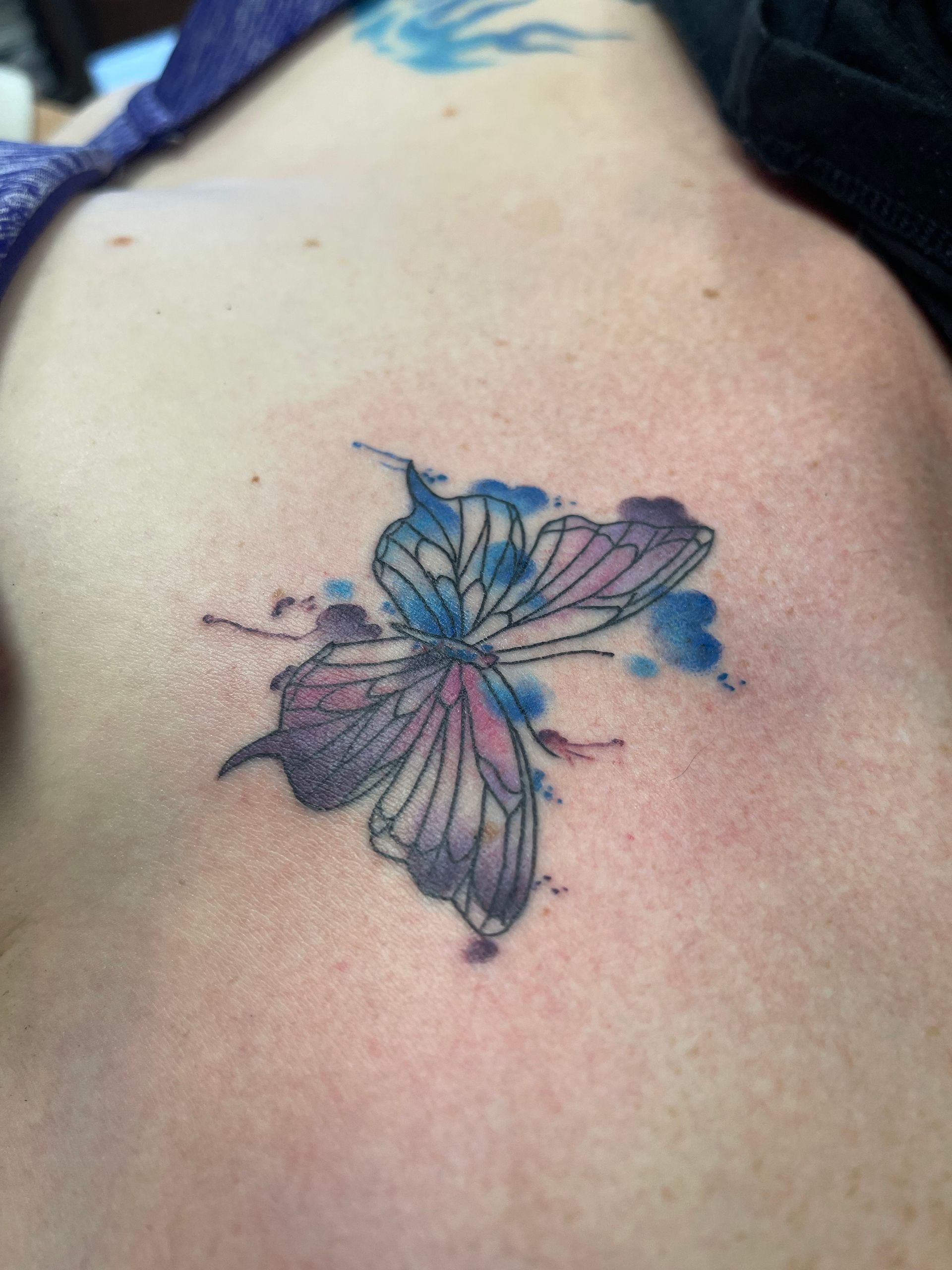 Watercolor butterfly tattoo on skin, with blue, pink, and purple hues.