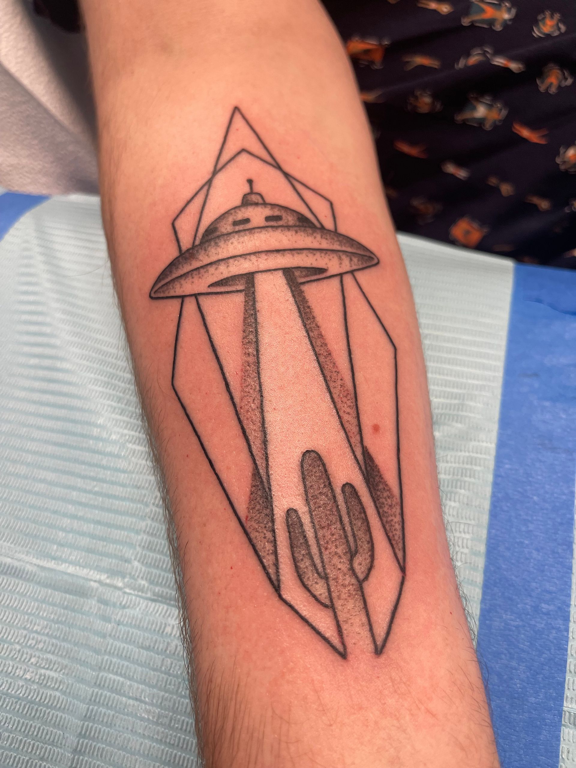 Tattoo on forearm: UFO beaming light onto cactus within a geometric diamond frame.