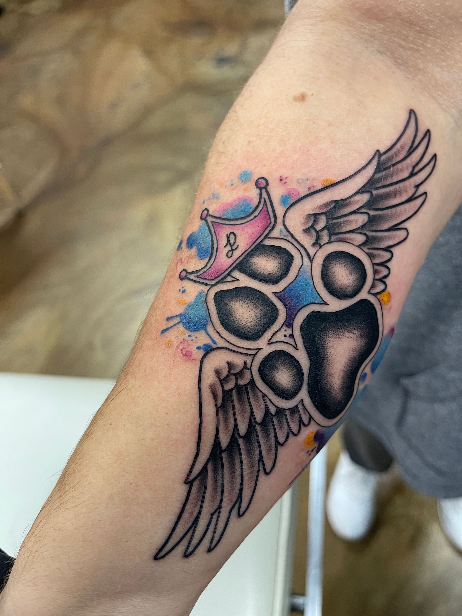 Tattoo of a dog paw print with wings and a crown on an arm. Features watercolor splashes.
