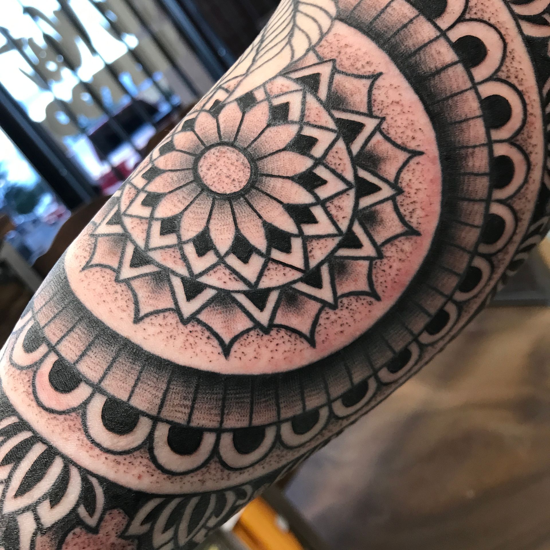 Arm tattoo with a black and gray mandala design featuring concentric circles and geometric patterns.