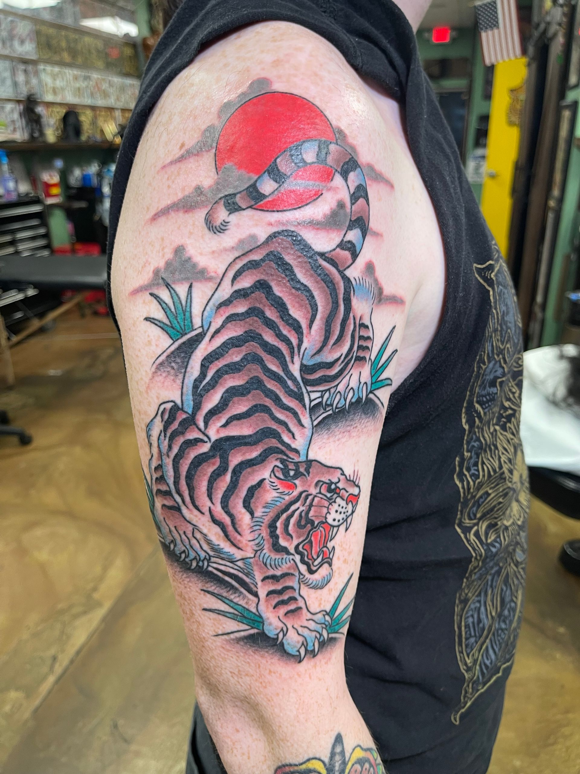 Tiger tattoo on a person's upper arm, with a red sun, grass, and a tank top.