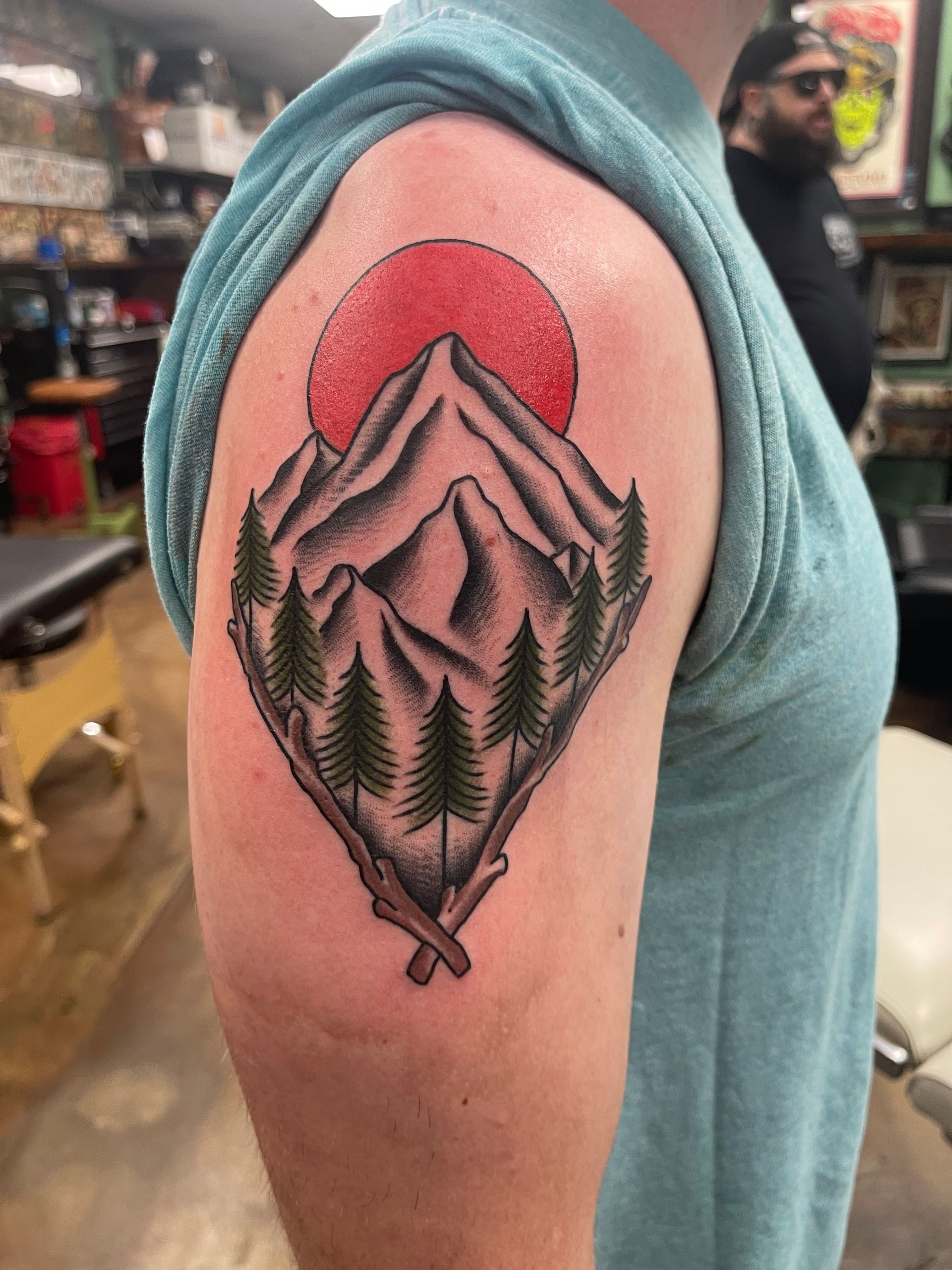 Tattoo of mountains, red sun, and trees on a person's upper arm.