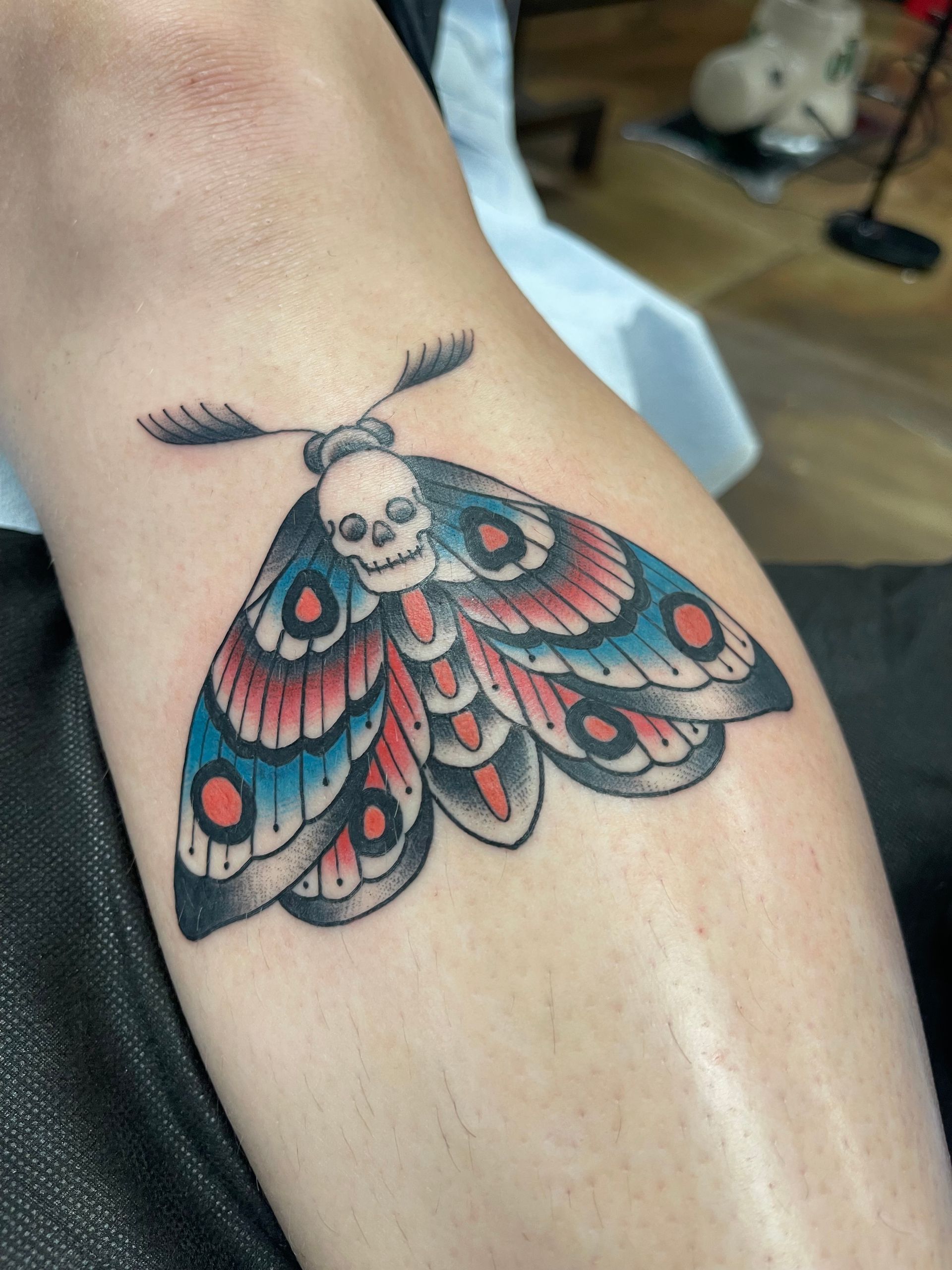 Tattoo of a moth with a skull head and blue, red, and black wings on a leg.