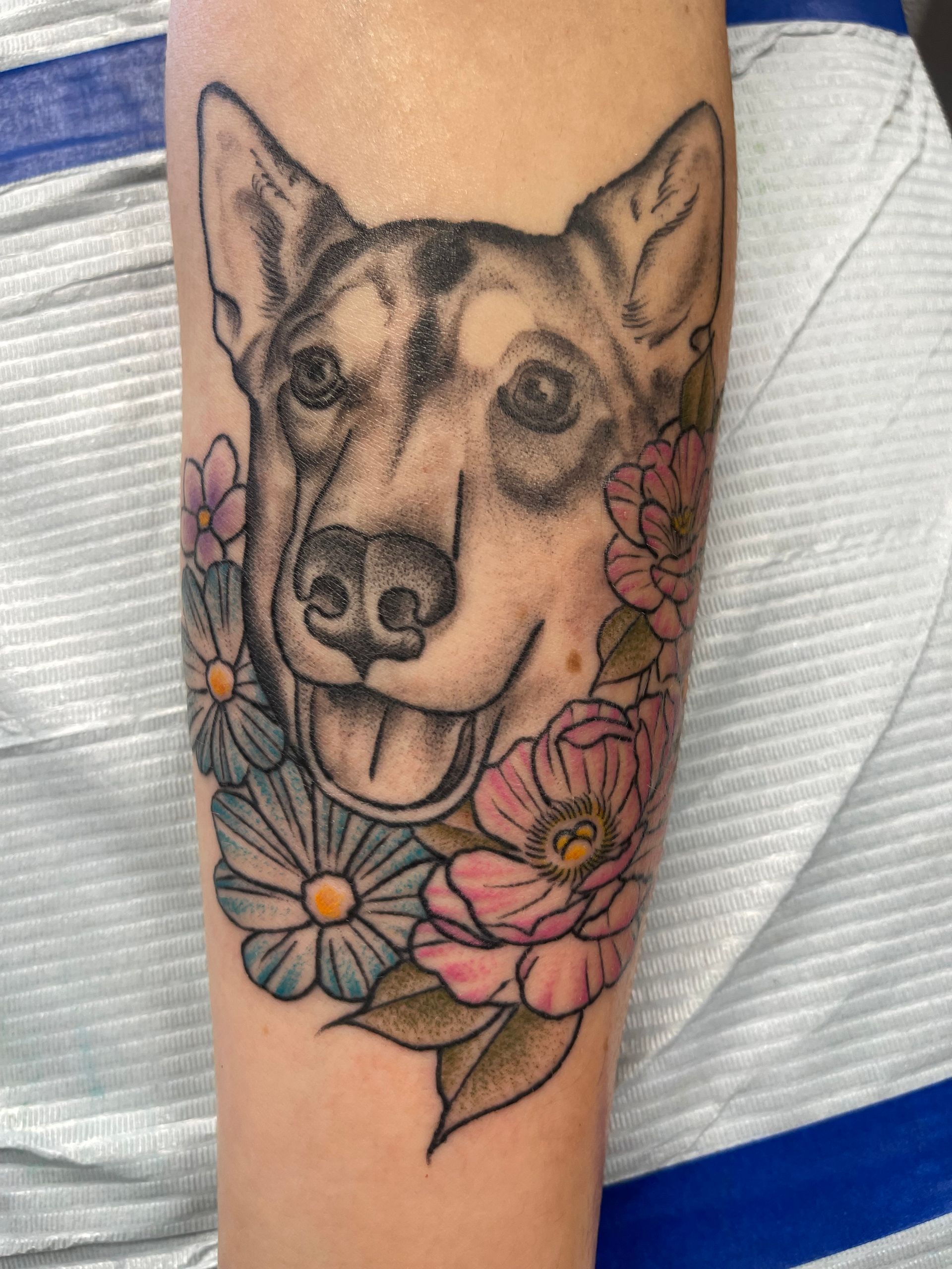 Tattoo of a dog's face with flowers; grayscale with pink, blue, and yellow on an arm.