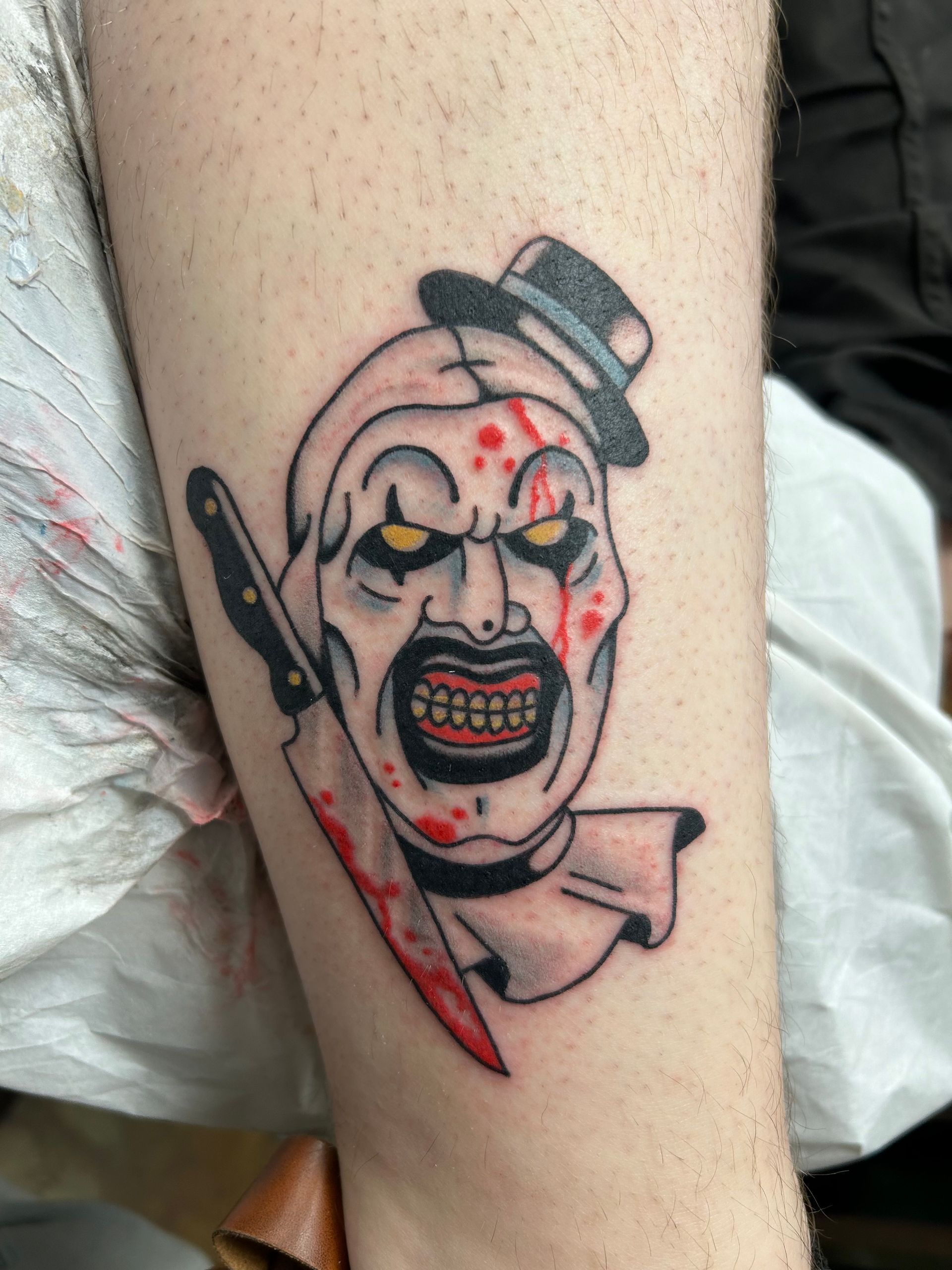 Tattoo of a scary clown's face with a bloody knife. White skin, red blood, and a small black hat.