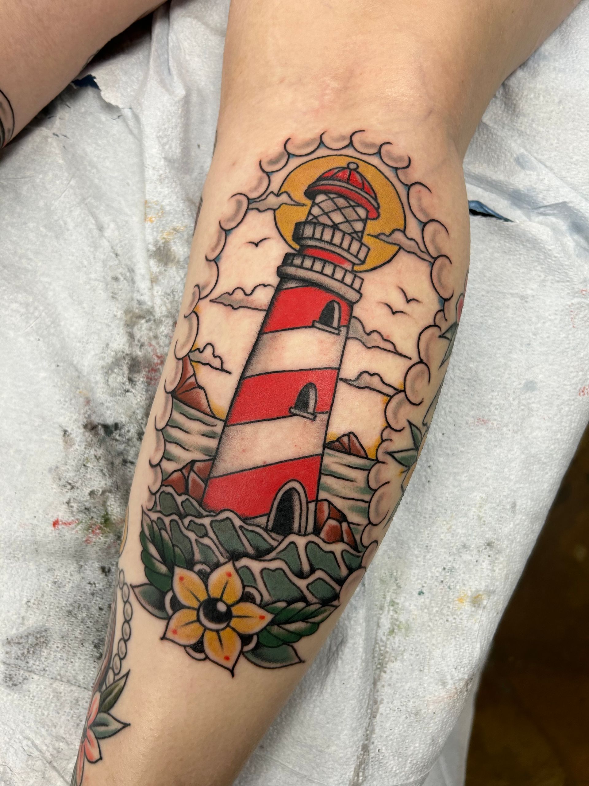 Tattoo of a red and white lighthouse, surrounded by clouds and ocean waves, on a person's leg.