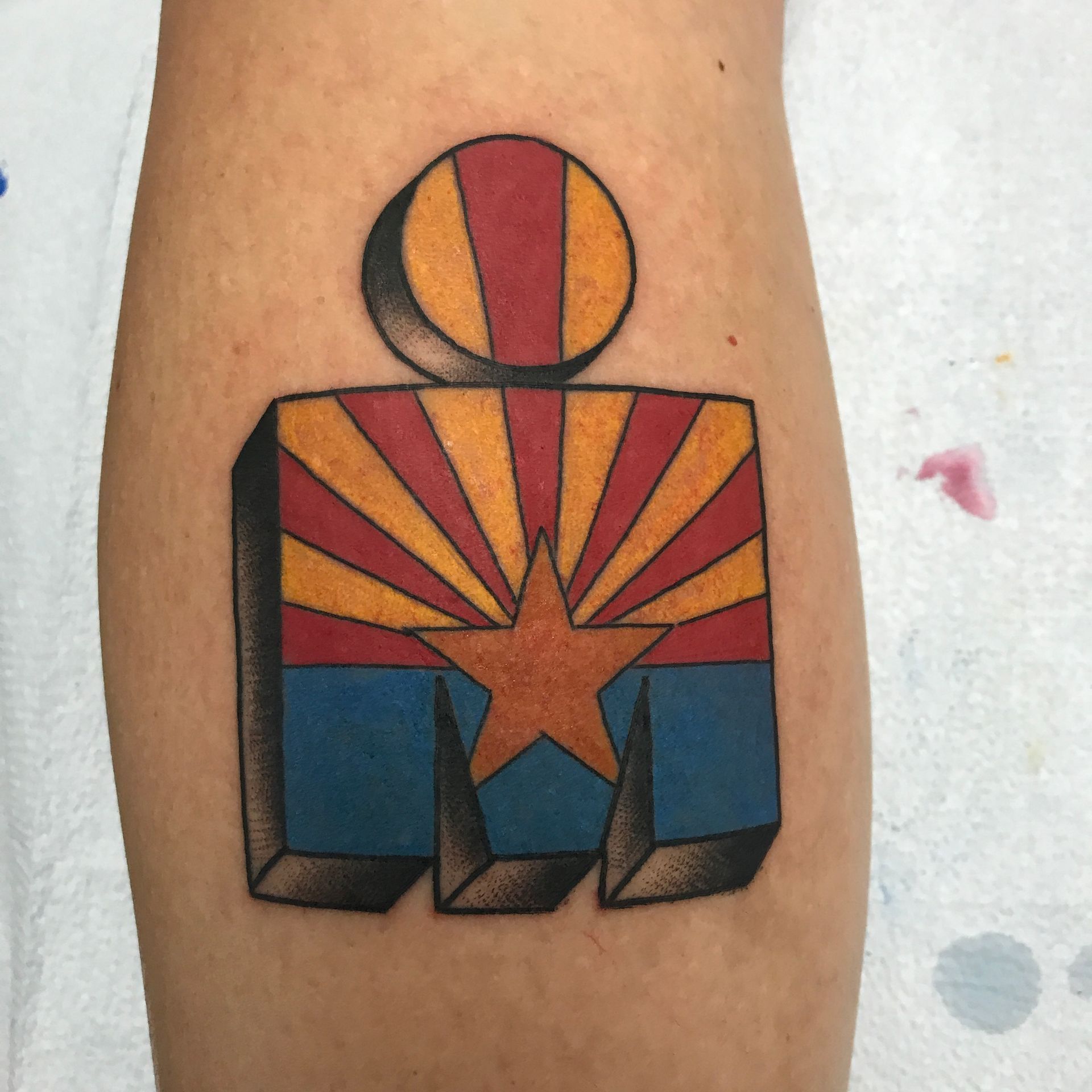 Tattoo on leg of Ironman logo stylized with Arizona flag colors, including a sunburst and star.