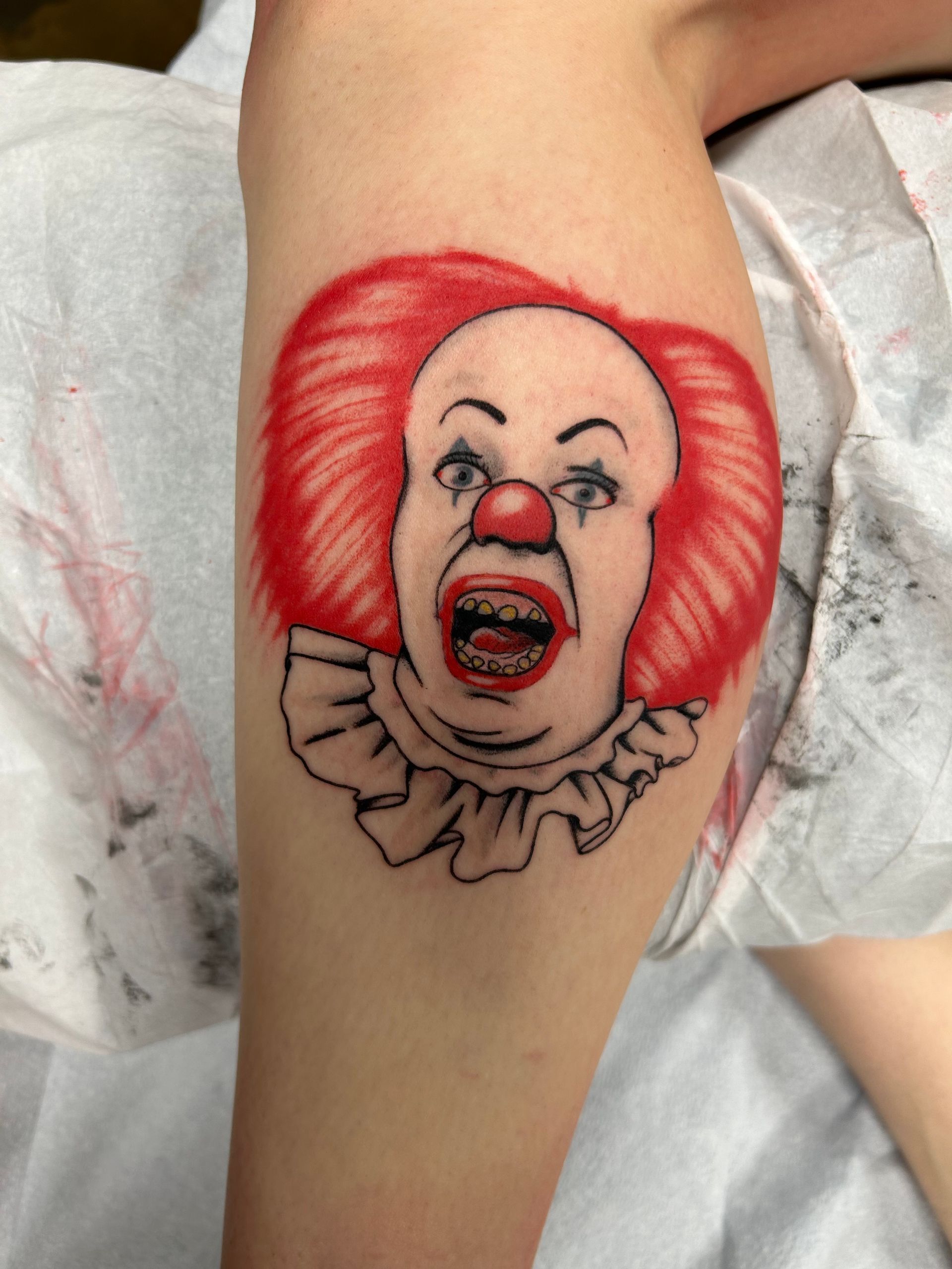 Tattoo of Pennywise the clown on a leg, red hair and nose, open mouth, white ruff.