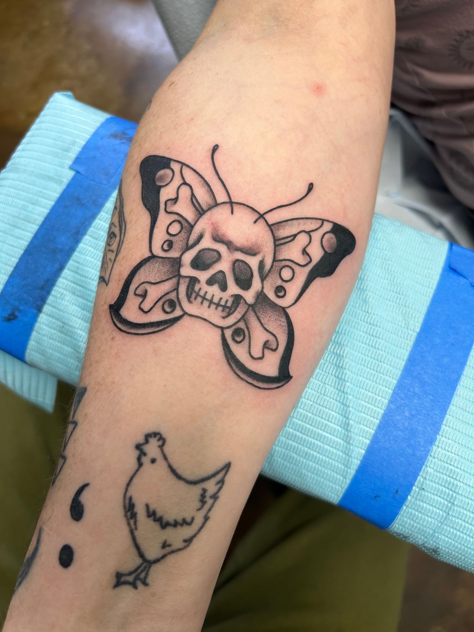 Tattoo on arm: skull butterfly, with a chicken and semicolon tattoo.