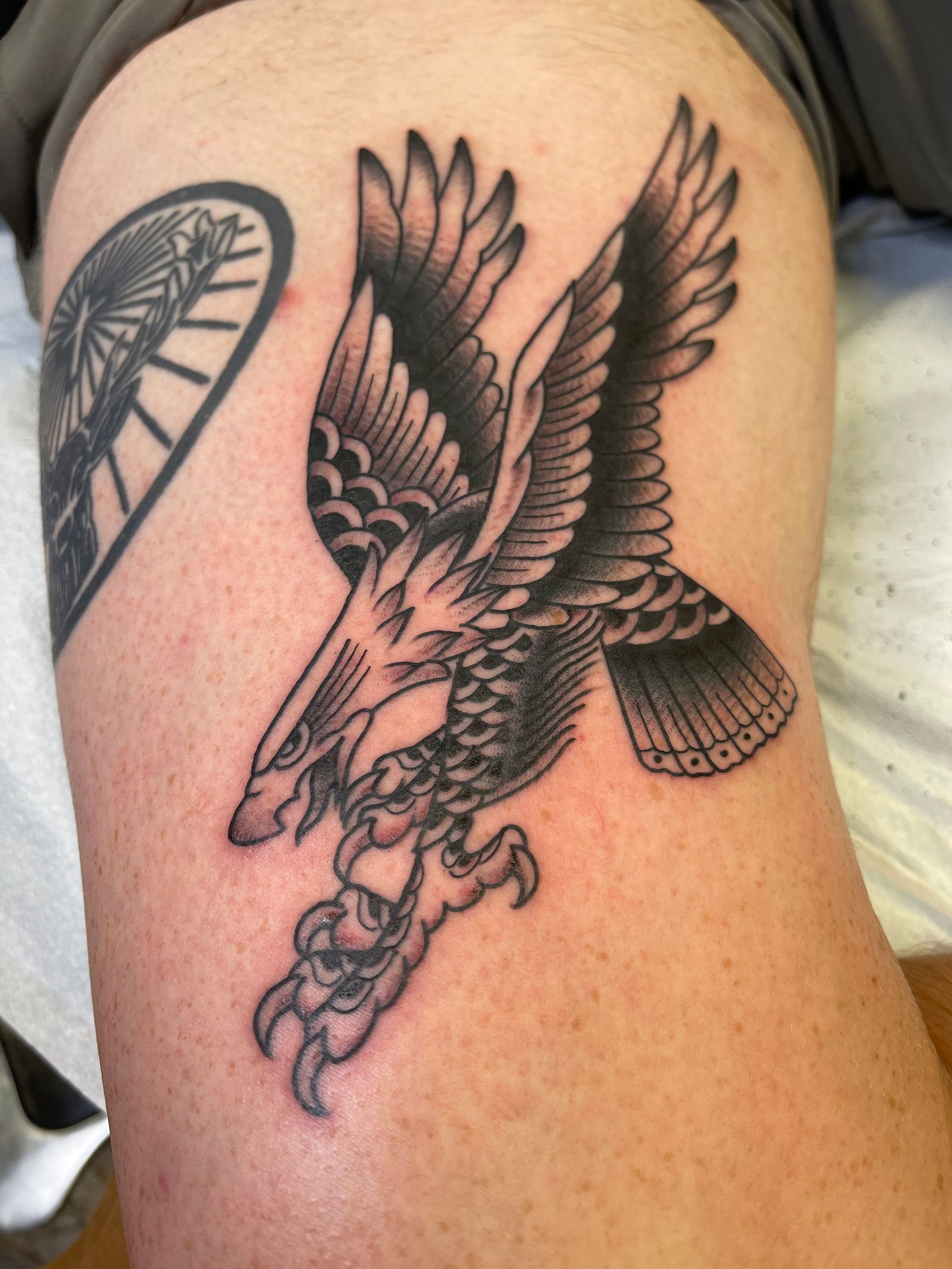 Black and gray eagle tattoo on a thigh, wings outstretched, talons extended.